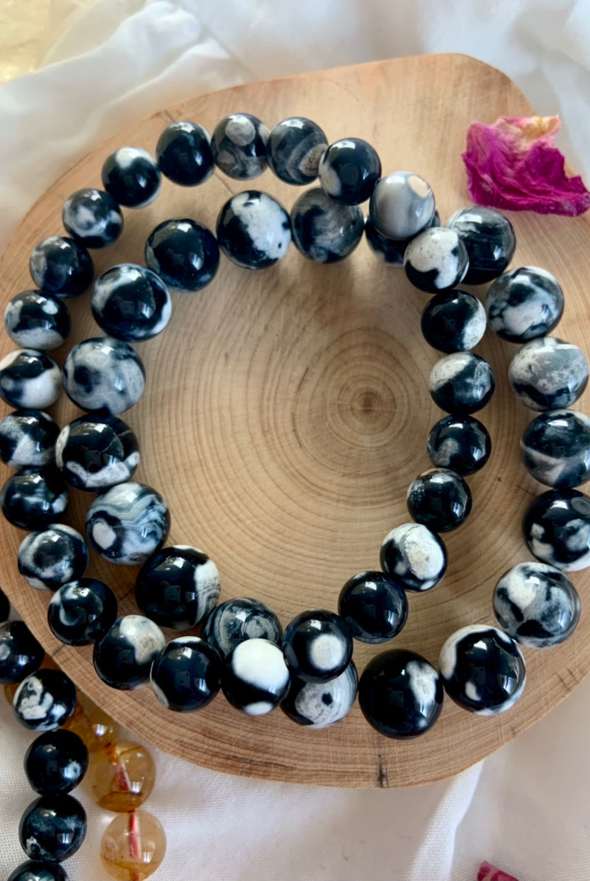 Overhead view of Orca Agate bracelets in 10–11mm and 8–9mm bead sizes resting on a natural wood slice with a pink rose petal for a soft, giftable vibe