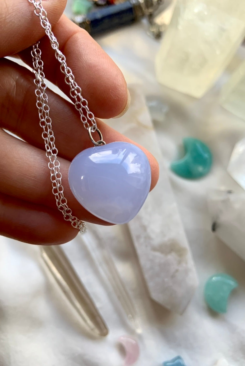 A luminous blue chalcedony puffy heart pendant necklace on s925 silver chain makes a sweet gift her. This pastel blue crystal resonates with the throat + heart chakras. Shown on a hand for scale, with crystals in the background.