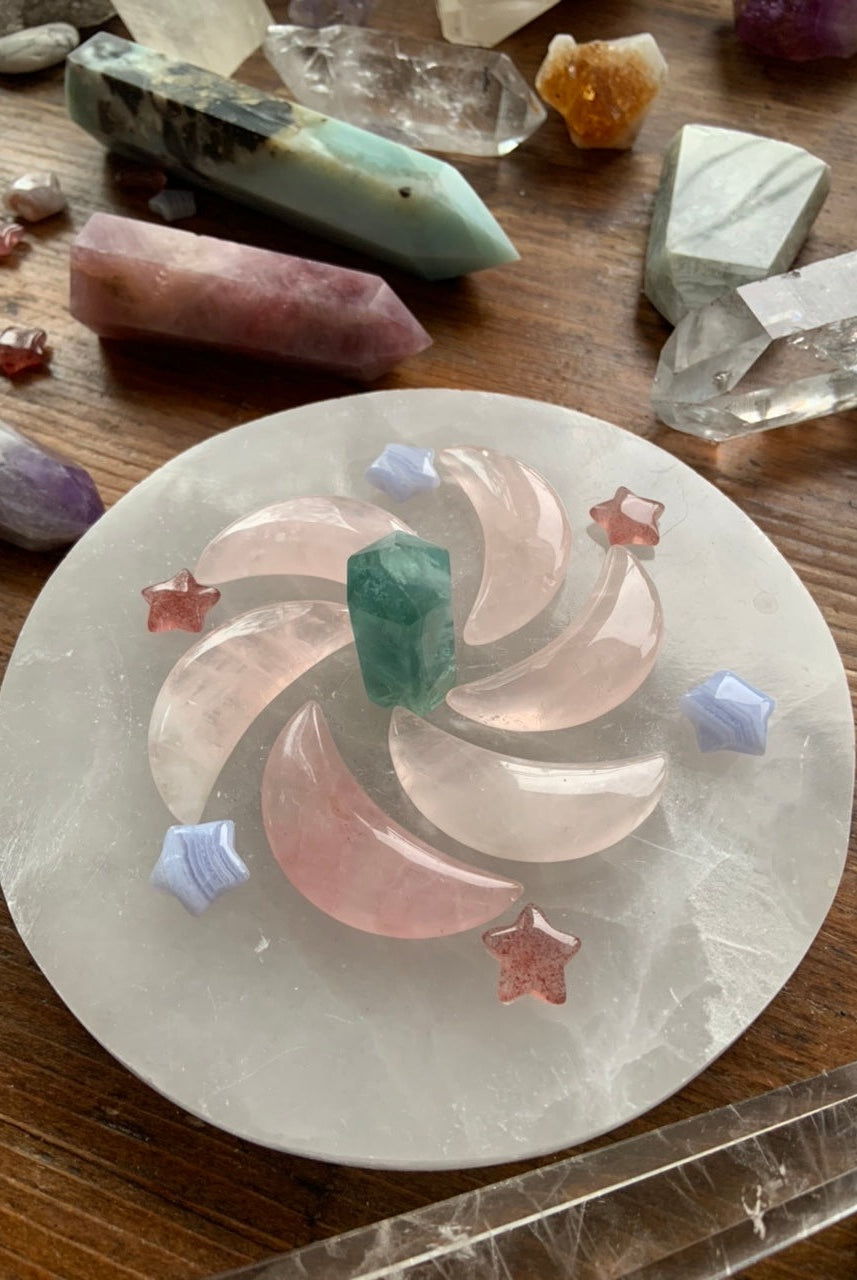 Crystal grid spiral of rose quartz moons, blue lace agate stars, and strawberry quartz stars on selenite plate with other crystals on dark wood table