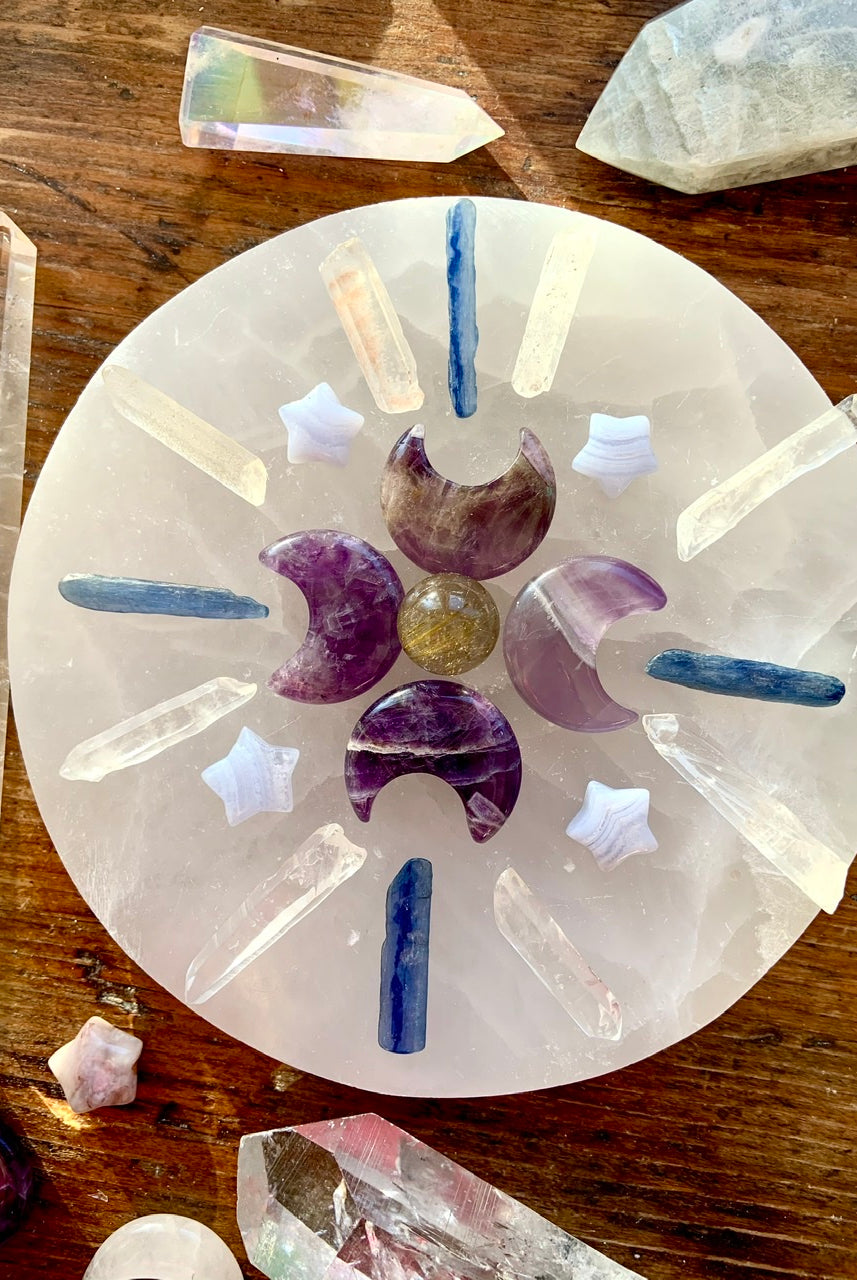 Overhead of crystal grid on selenite plate with kyanite sticks, clear quartz points, amethyst moons, and blue lace stars on dark wood altar table