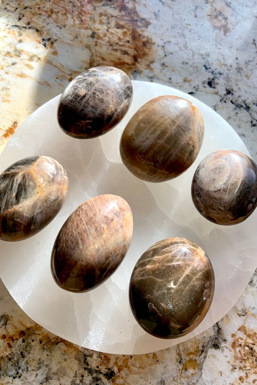 An array of Black Moonstone Palm Stones shown on a white selenite charging plate - crystals for empaths