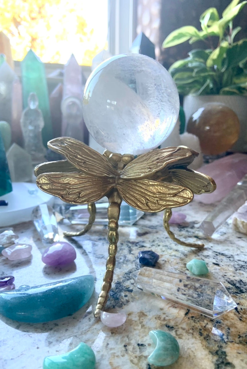 Gold dragonfly crystal ball stand holding a clear quartz crystal sphere, displayed in a colorful crystal collection with plants.