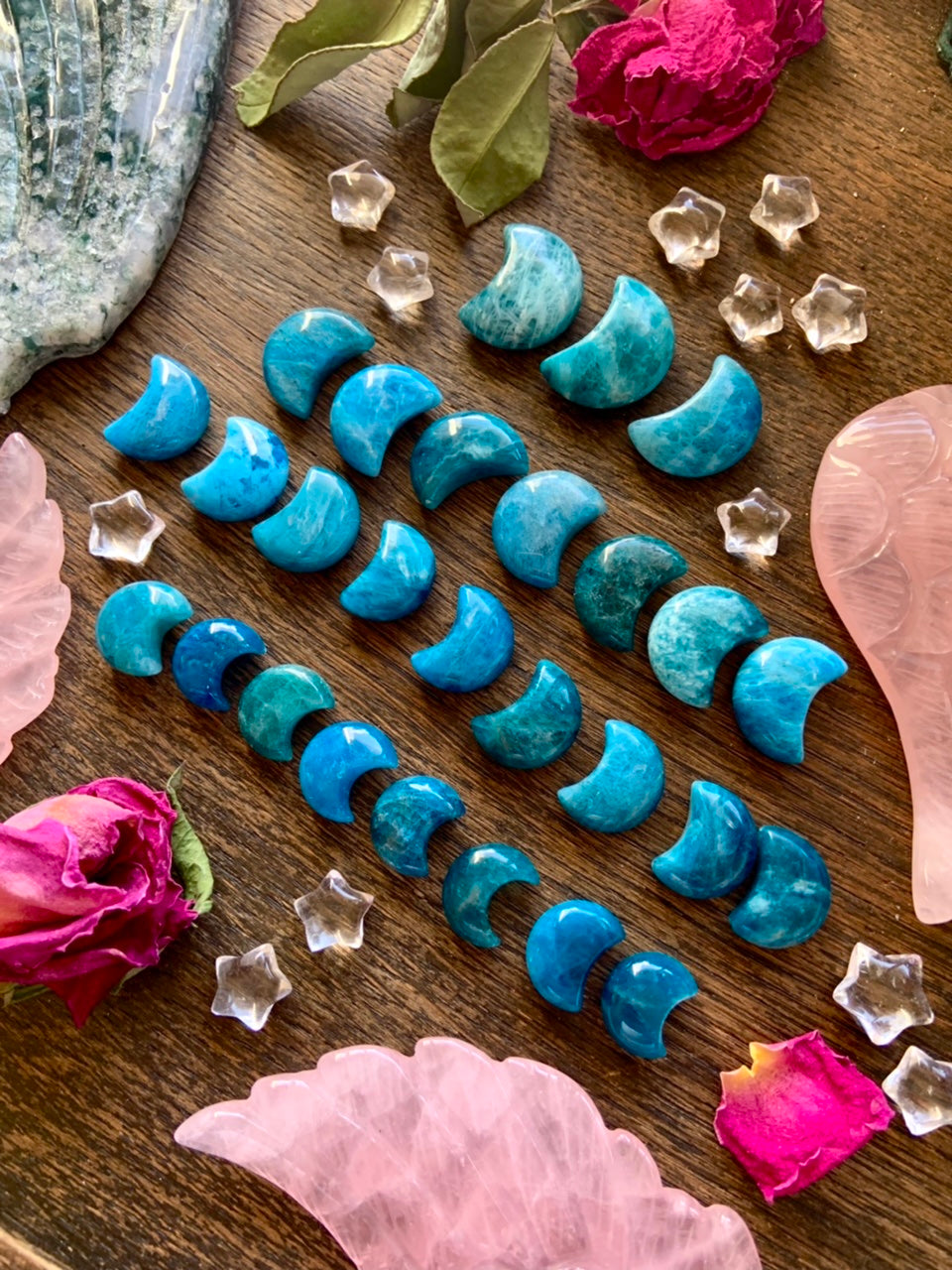 Wide diagonal array of Blue Apatite Crescent Moons on wood table — arranged by size with rose quartz angel wings, clear quartz stars & petals.