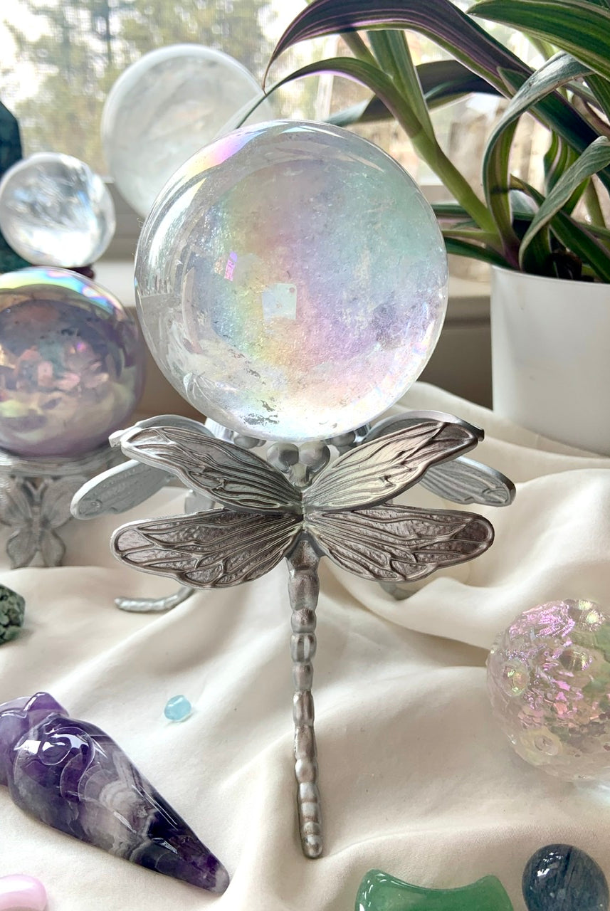 Crystal sphere stand with dragonfly design in silver, holding an aura quartz crystal ball in a dreamy display with plants & backlit crystals.