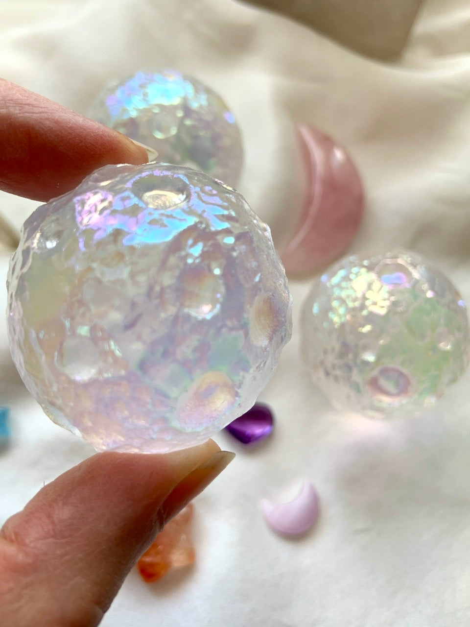 Angled close-up of Angel Aura Quartz moon sphere with surrounding crystals on white silk — celestial carving for divine insight & spiritual energy.