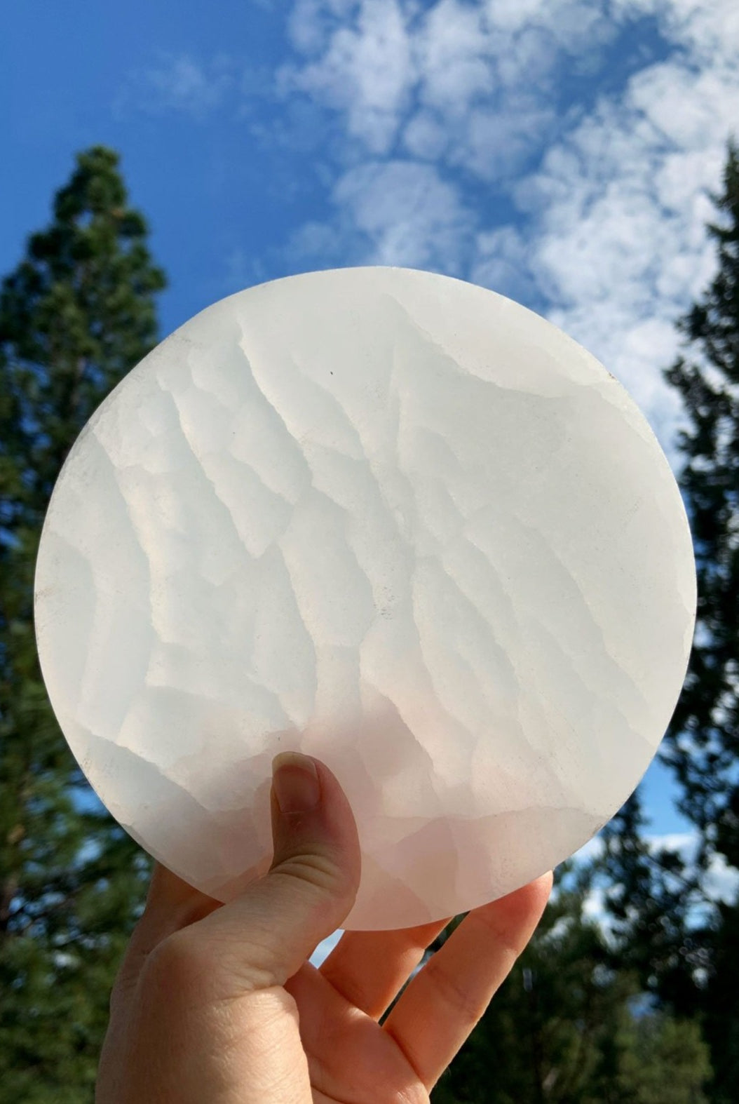Selenite charging plate held to the light outdoors with blue sky, white clouds, and tree silhouettes