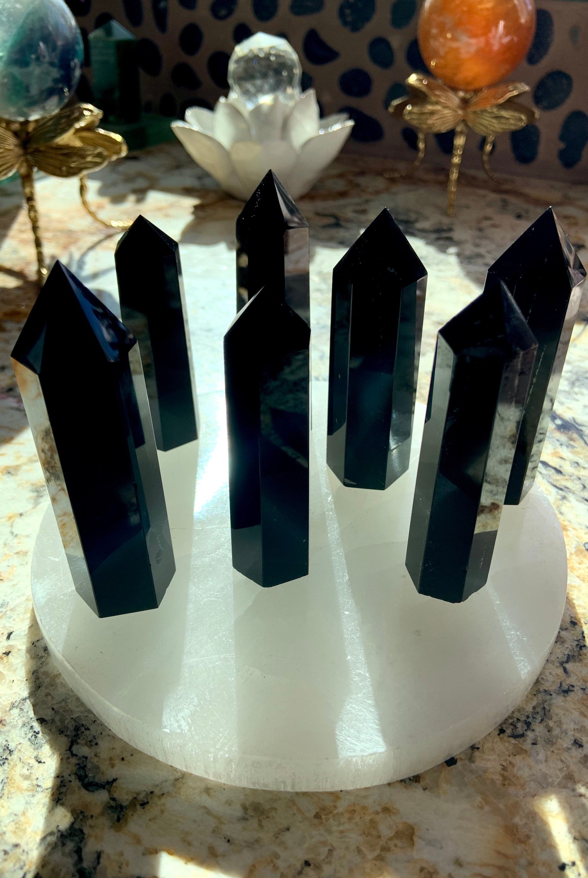 An array of black obsidian points shown in a group on top of a selenite plate with crystal spheres in the background. Choose your size from the dropdown menu.