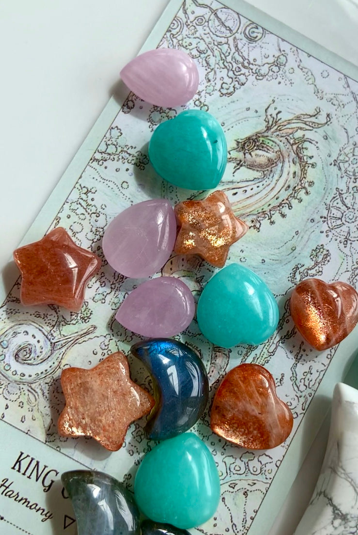 Mini crystal carvings including Sunstone stars, Amazonite hearts, Labradorite moons, & Kunzite teardrops arranged on a tarot card — crystals for healing, intention & energy work.