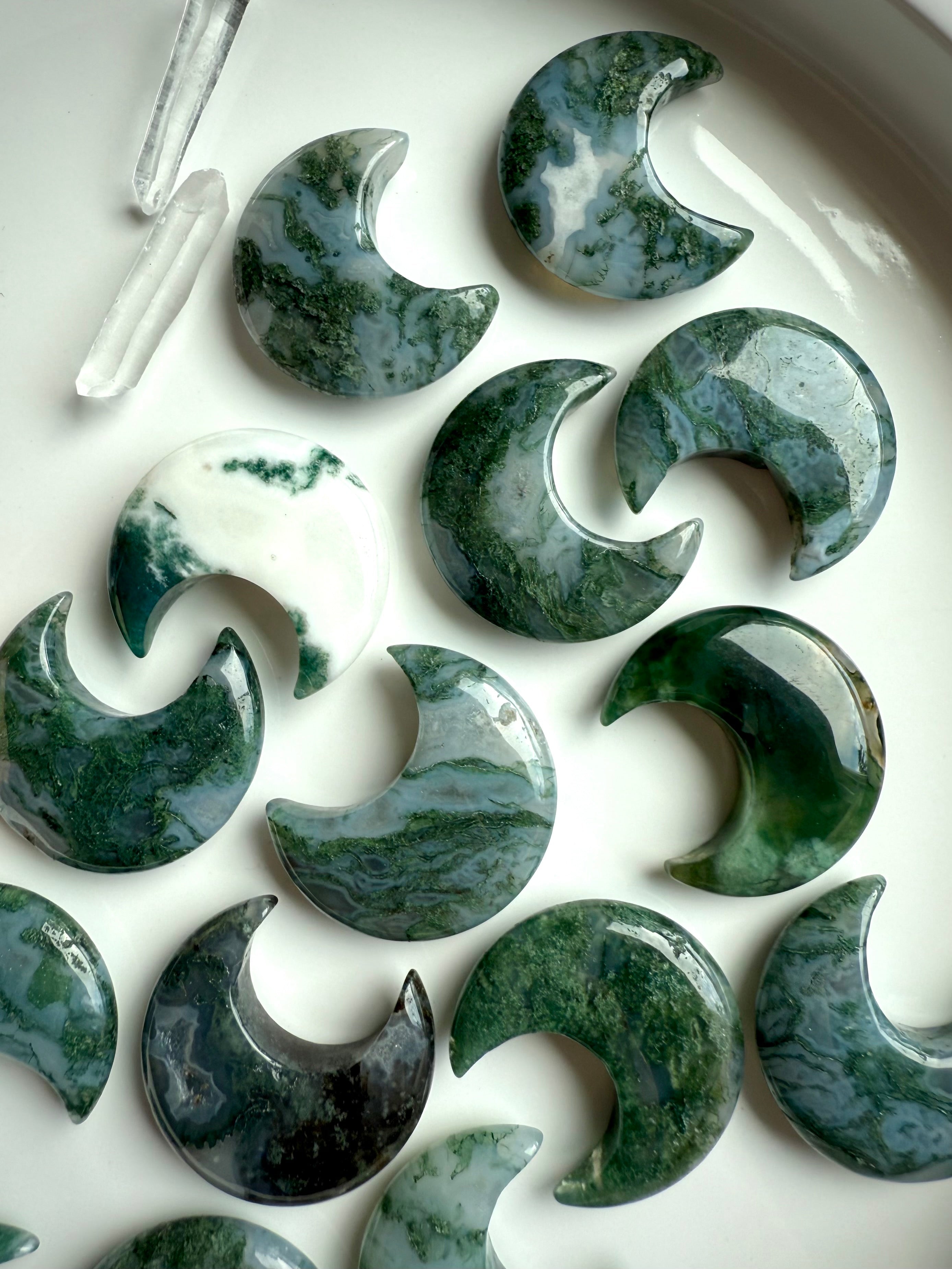 Overhead view of polished Moss Agate crescent moon carvings on a white plate, showcasing swirling green and white patterns with Clear Quartz points.