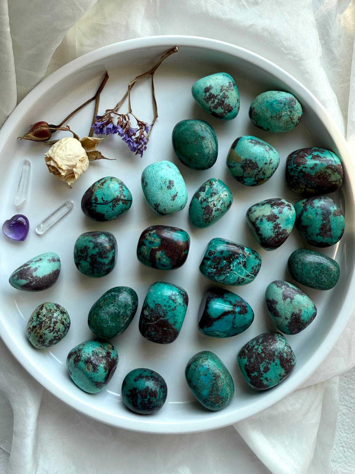 Polished Chrysocolla tumbled stones with blue-green tones and black matrix, styled in a white dish with dried flowers and crystals for heart and throat chakra healing.