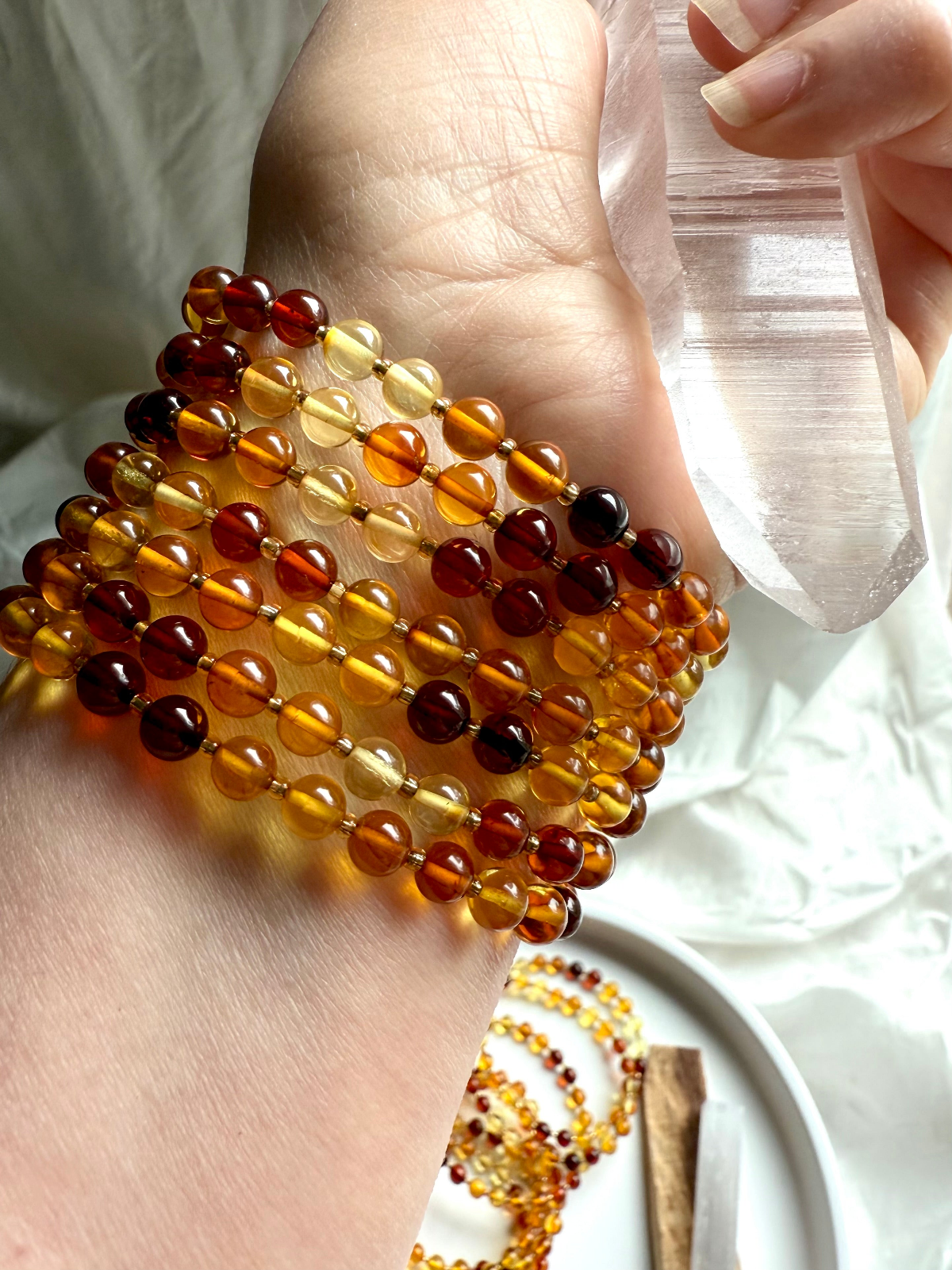 Stack of radiant Baltic Amber bracelets in golden, honey, and cherry hues wrapped around a wrist holding a clear quartz point — vibrant crystal jewelry for Solar Plexus chakra healing, energy, and natural beauty.