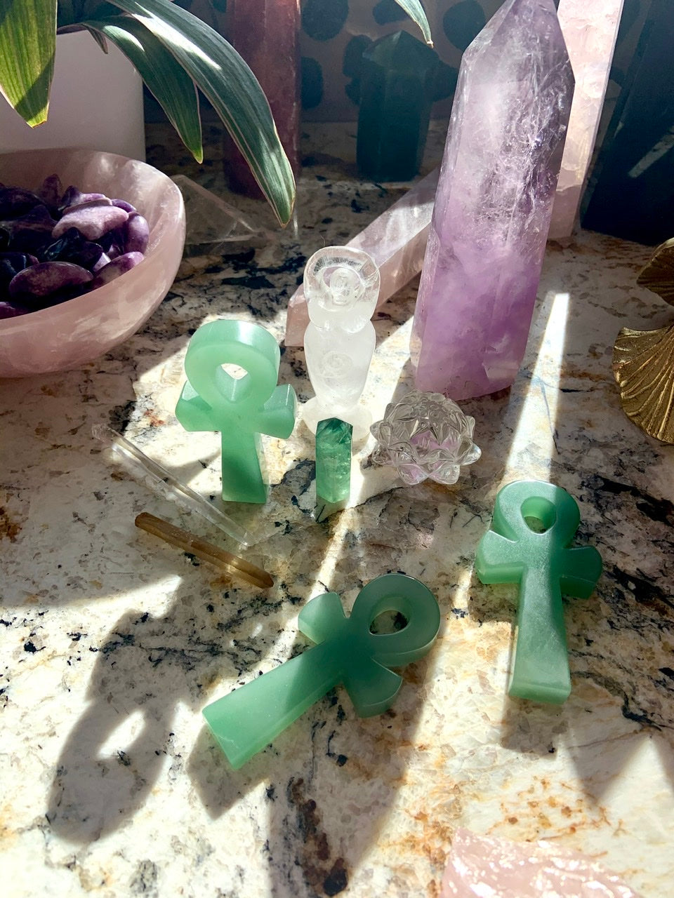 Green Aventurine ankh crystal carvings with sunlight pouring in, alongside a Clear Quartz goddess, citrine and fluorite points, and a lotus carving — mystical crystal altar arrangement.