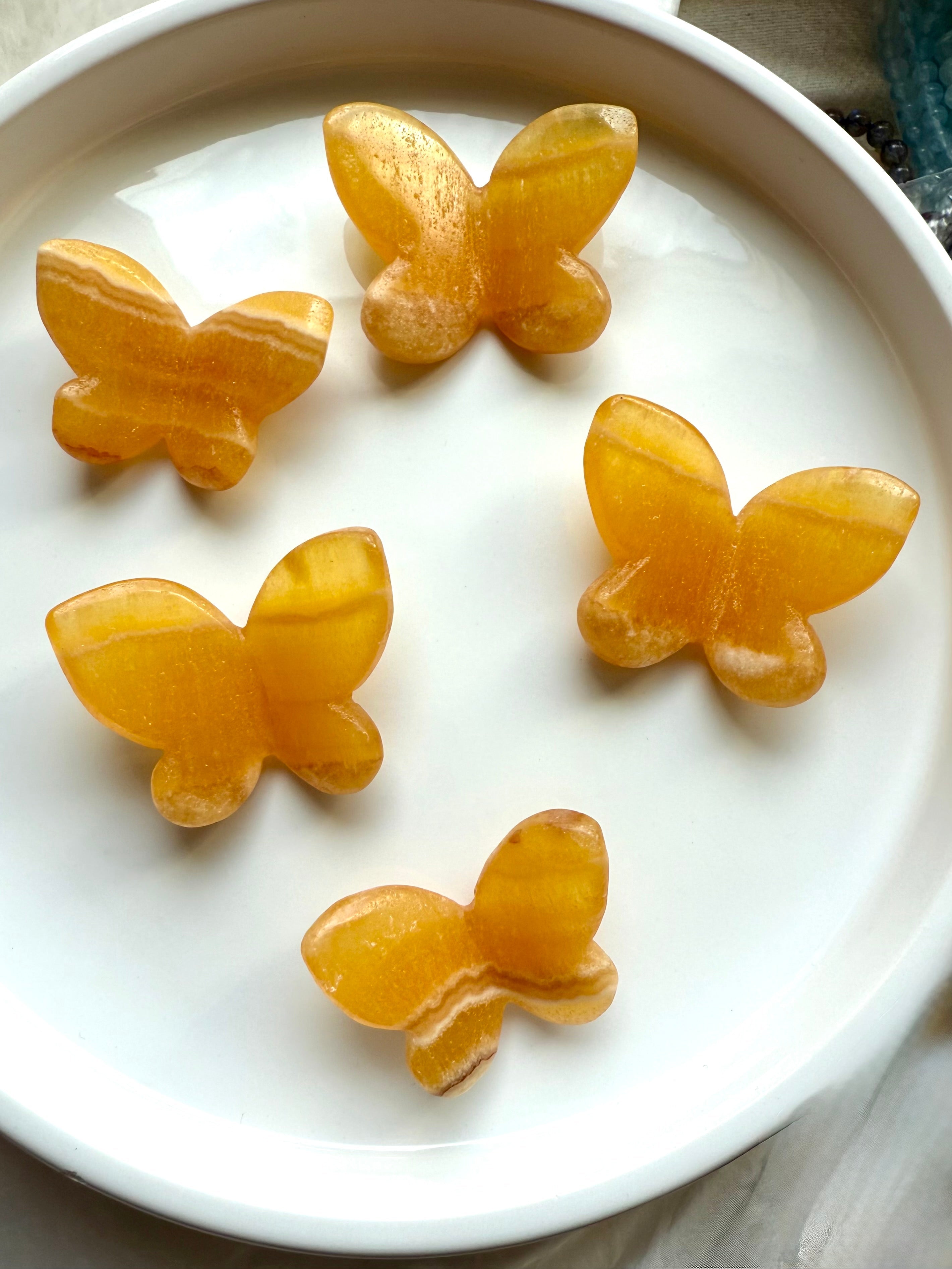 Orange Calcite butterfly crystal carvings on a white dish, glowing with vibrant golden-orange hues — symbolic of transformation, creativity, and solar plexus chakra activation.