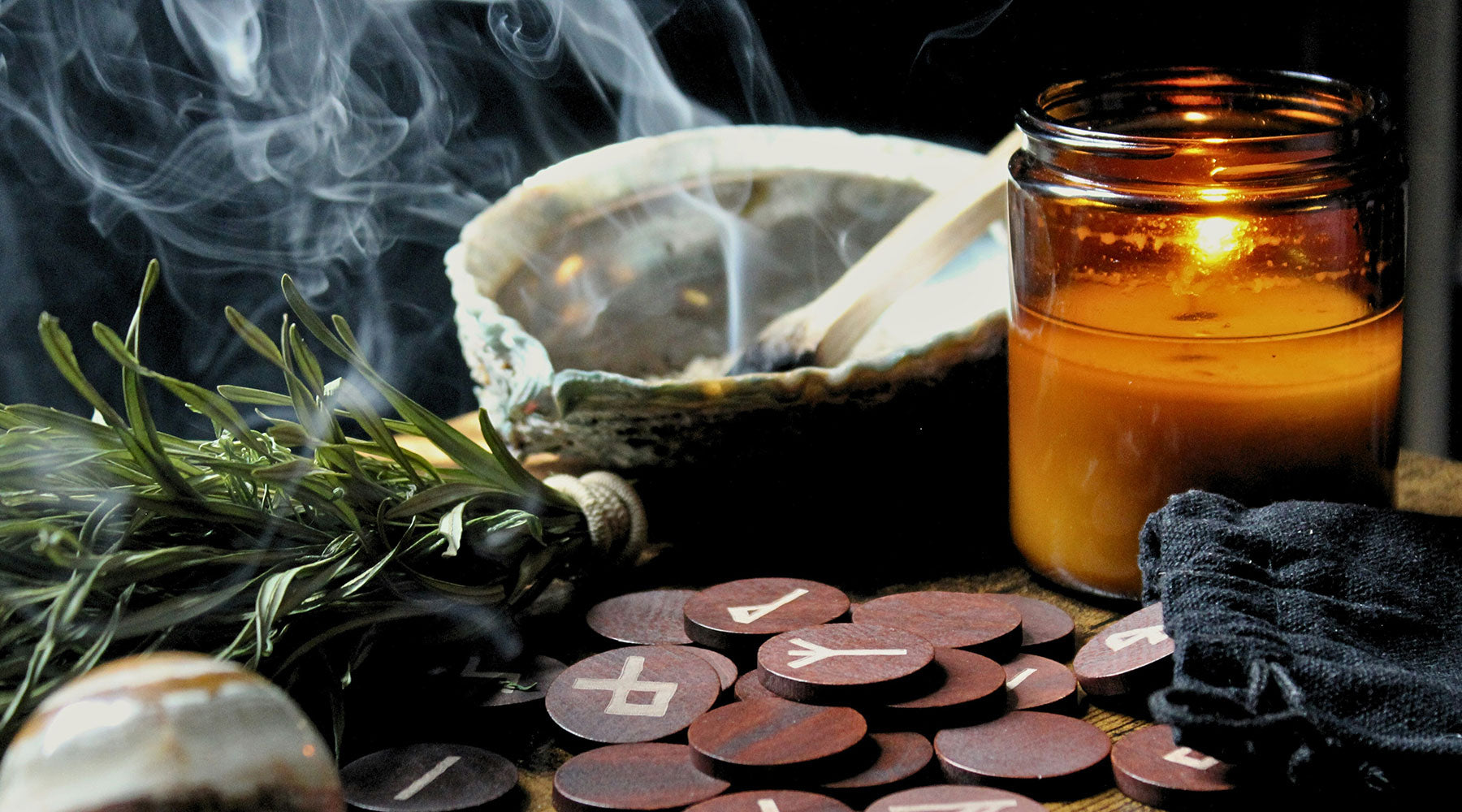 Rosemary smudge bundle with palo santo smoke rising from an abalone shell, shown alongside a lit candle, crystal ball, and rune stones — sacred tools for energy clearing rituals.