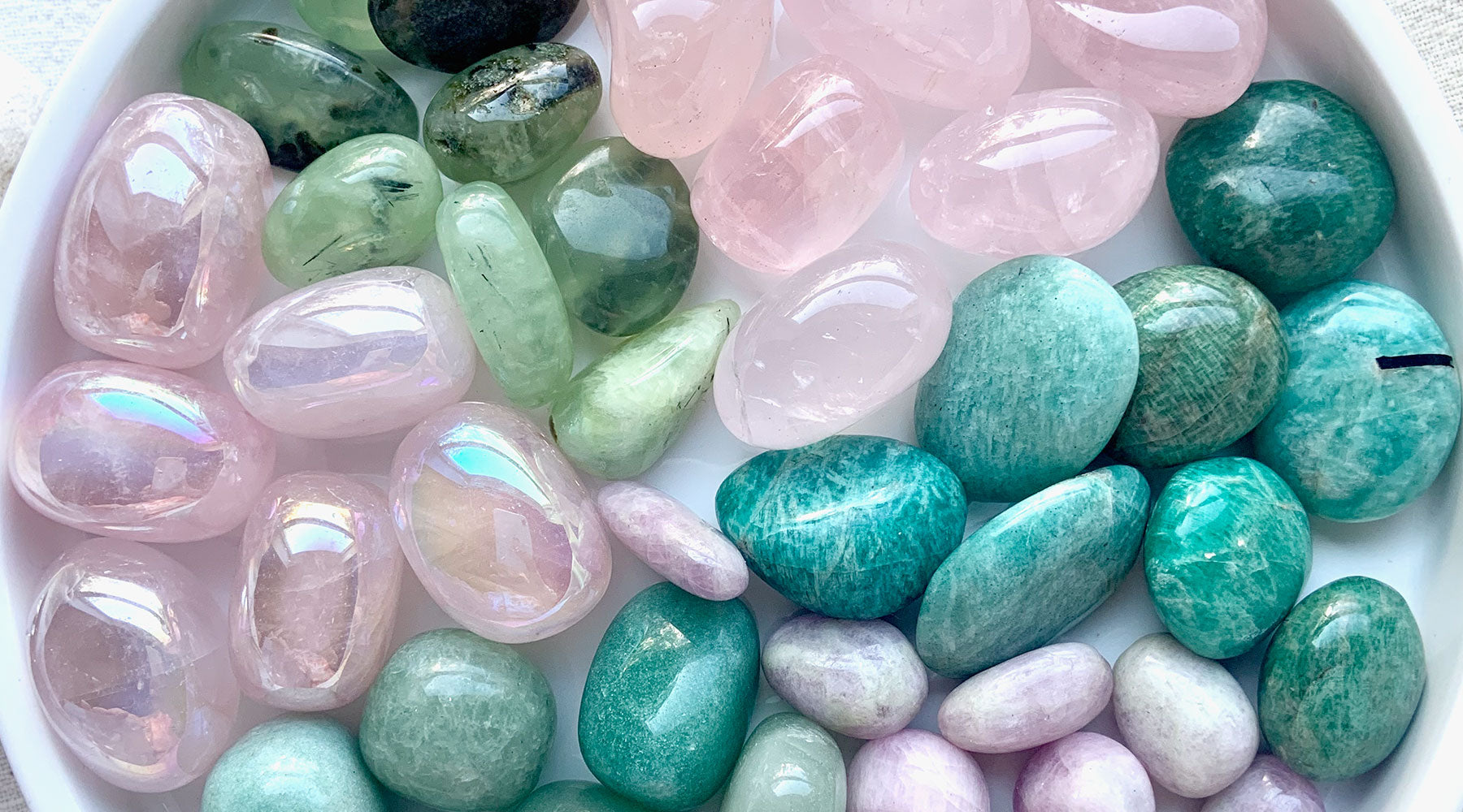 A colorful flatlay of pink and green tumbled stones including Rose Quartz, Green Aventurine, Kunzite, Prehnite, Amazonite, and Aura Rose Quartz — heart chakra crystals for love, compassion, and emotional healing.