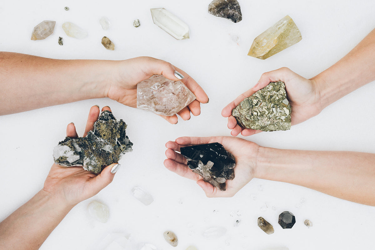 Flatlay of two women’s hands holding protective crystals including Pyrite, Smoky Quartz, and Clear Quartz, with other stones arranged for energy shielding.