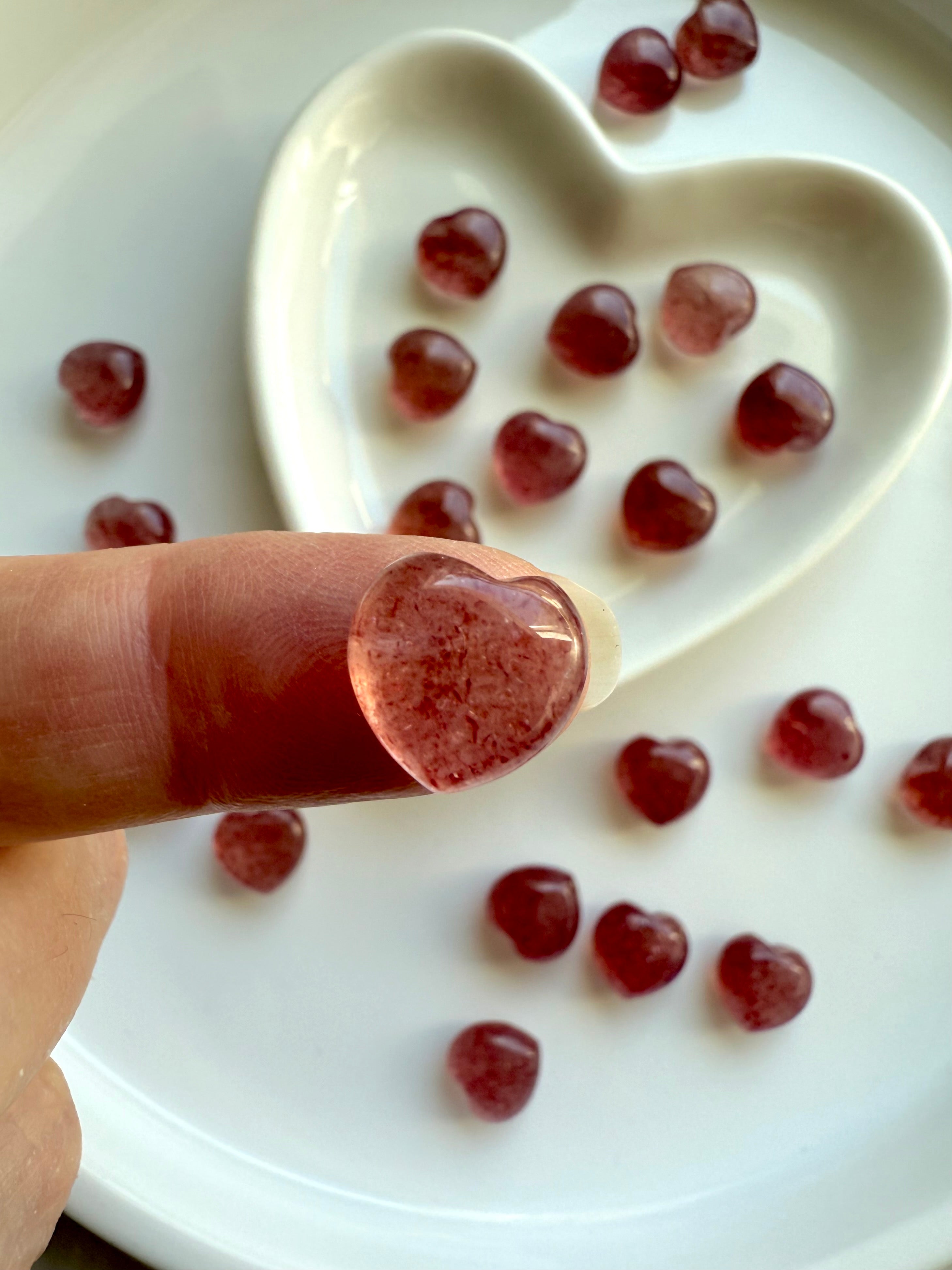 Tiny Strawberry Quartz heart held on fingertip above a ceramic bowl and plate of scattered hearts — showcases mini size and deep red to pink hues.