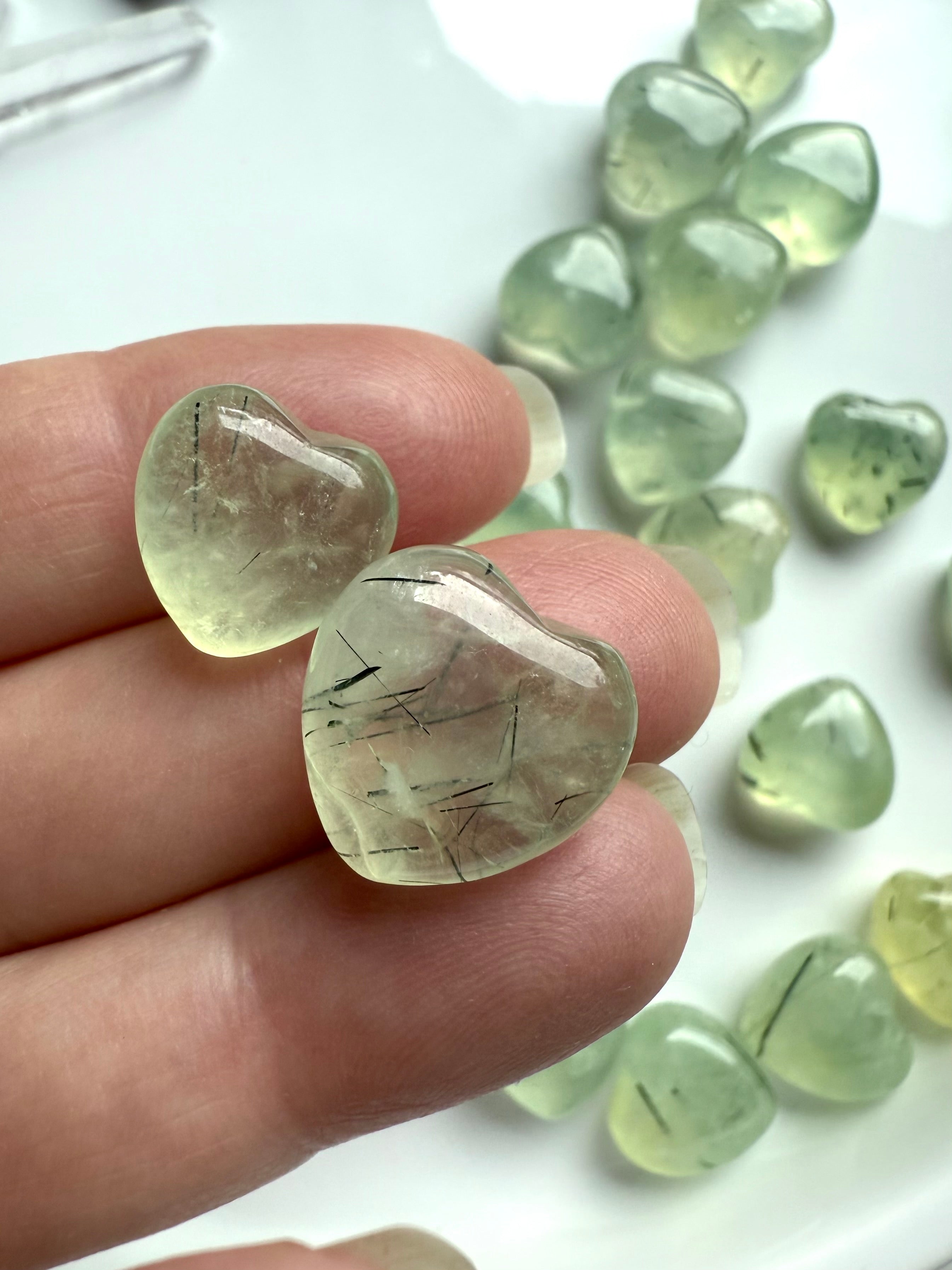 Close-up of two polished Prehnite crystal hearts with dark green inclusions, held in fingers for detail.