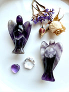 Dream Amethyst angel carvings with chevron banding, styled on a white plate with dried rose, purple statice flowers, Amethyst heart, and clear quartz gemstone – crystal angels for healing, grief, and spiritual protection