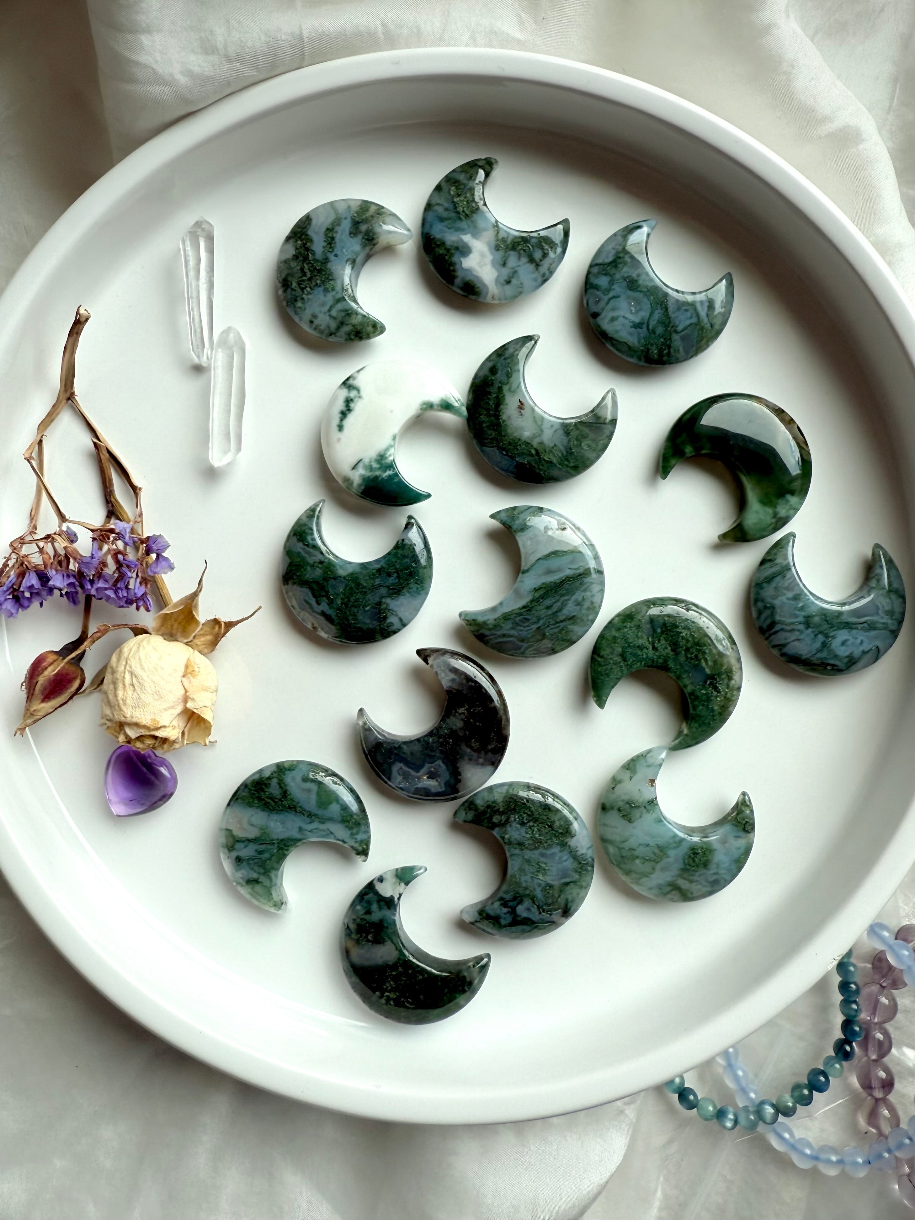 Moss Agate crescent moons with Clear Quartz points and dried flowers arranged on a white plate in a celestial altar flatlay.
