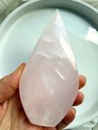 Mangano Calcite flame crystal held in hand for scale, featuring soft pink translucency and a smooth polished finish from Pakistan