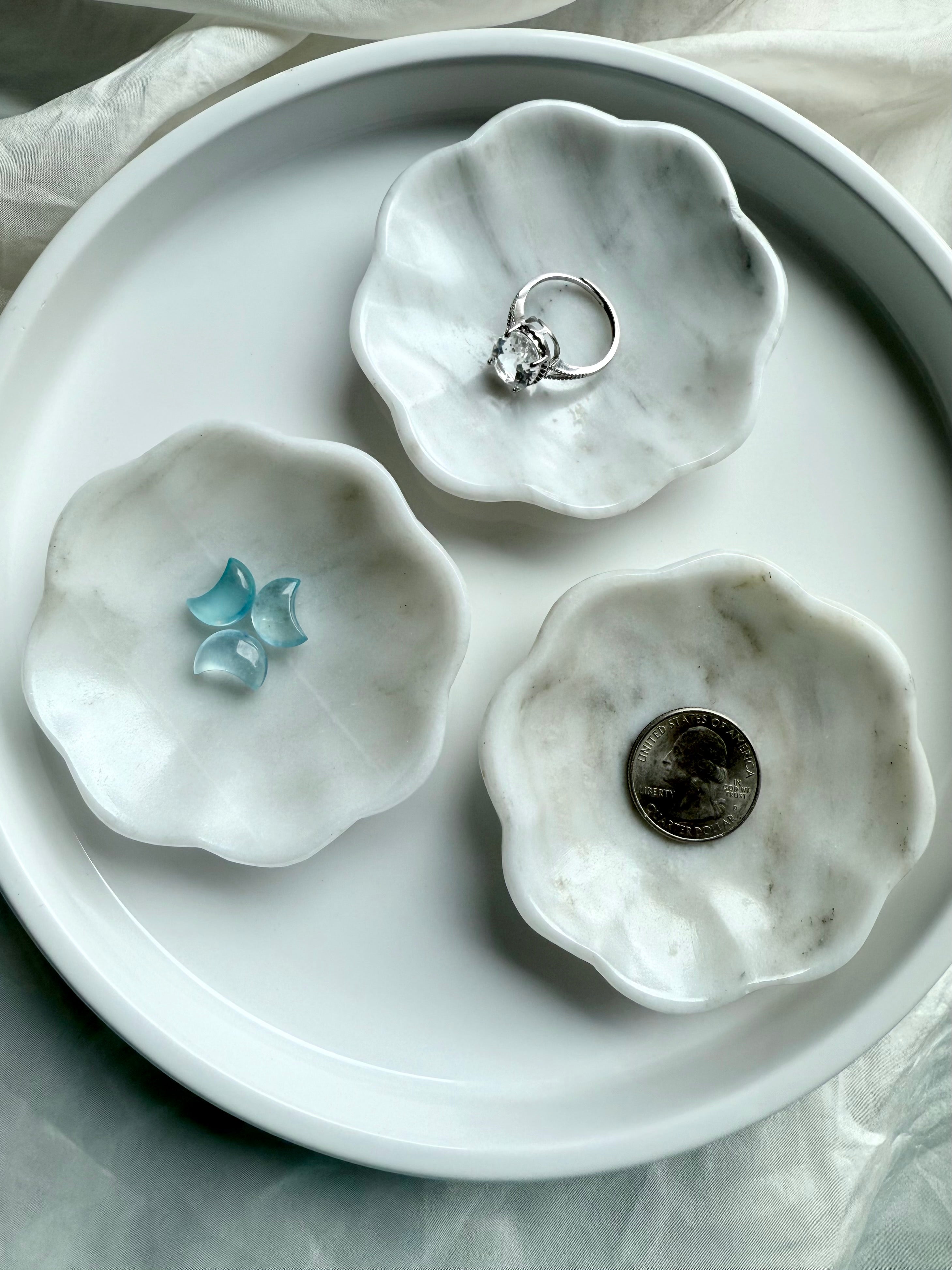 Three white marble daisy bowls with natural gray veining, one styled with a silver ring, one with aquamarine crescent carvings, and one with a quarter for size reference