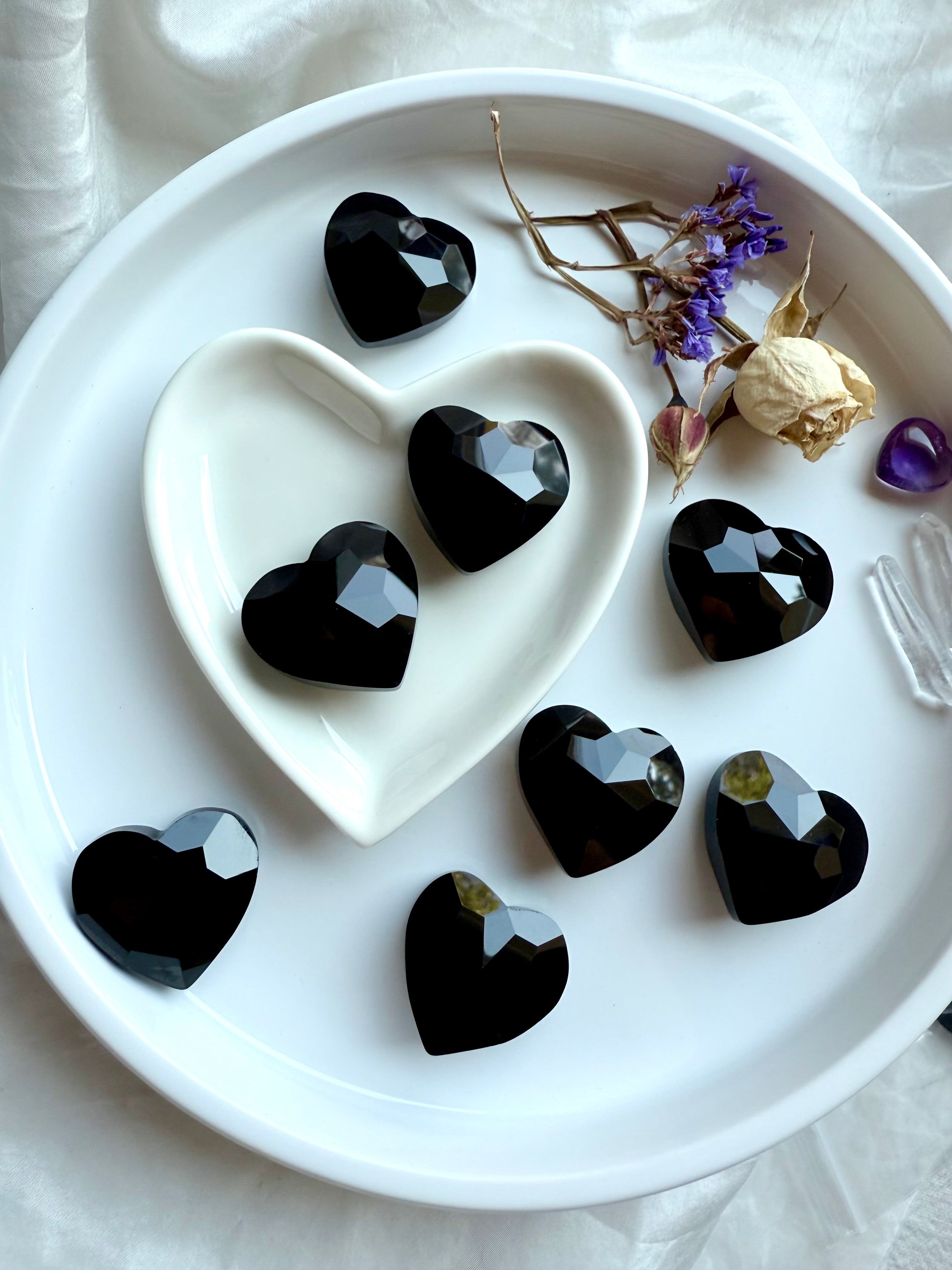 Faceted black obsidian heart-shaped stones in a white heart-shaped dish with crystals and dried flowers.