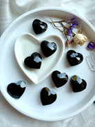 Faceted black obsidian heart-shaped stones in a white heart-shaped dish with crystals and dried flowers.