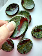 Dragon Blood Stone thumb stone held between fingers, revealing its curved indentation and striking red and green mineral patterns