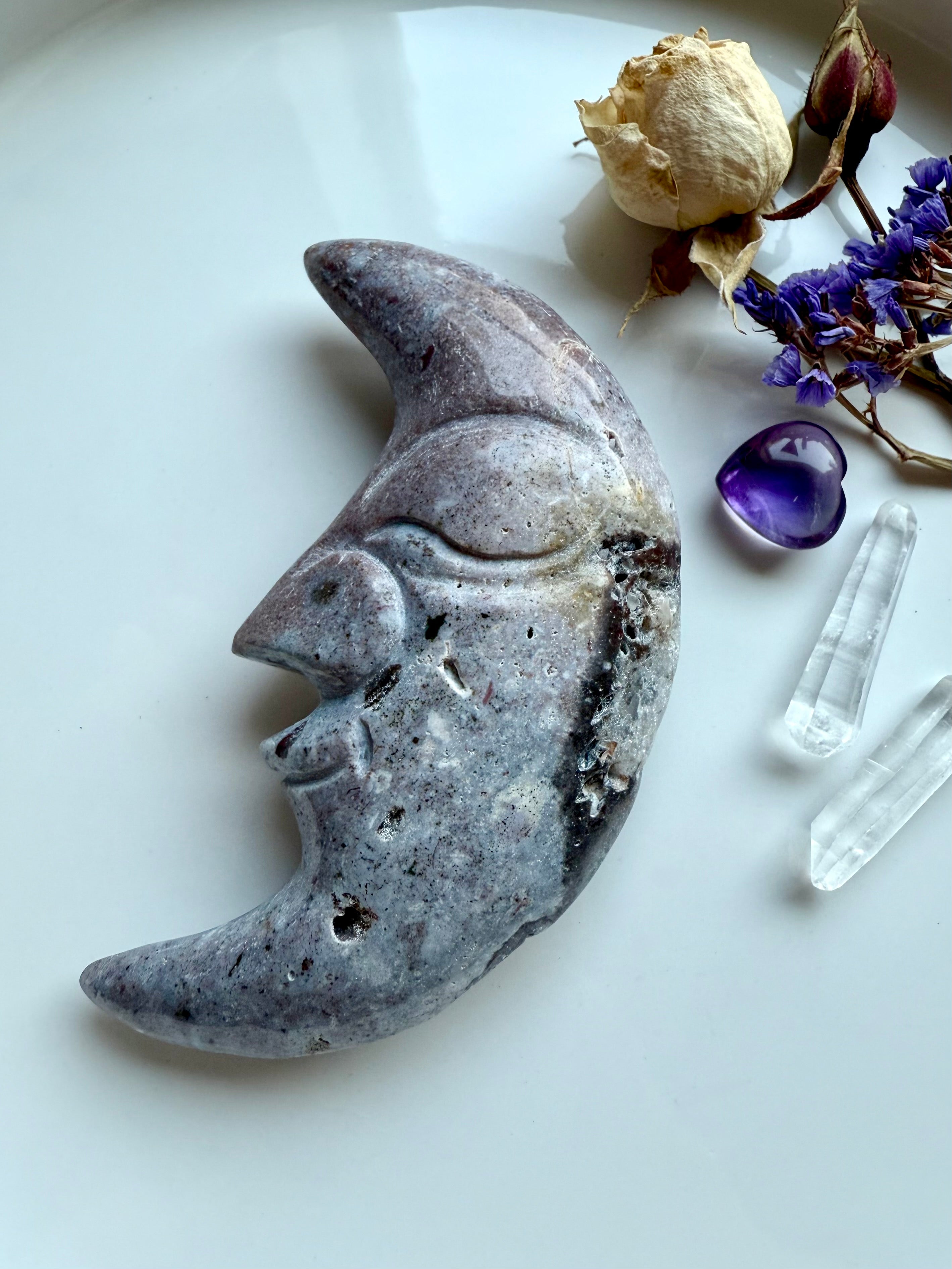 Purple Ocean Jasper moon face crystal carving displayed with quartz points, amethyst heart, and flowers.
