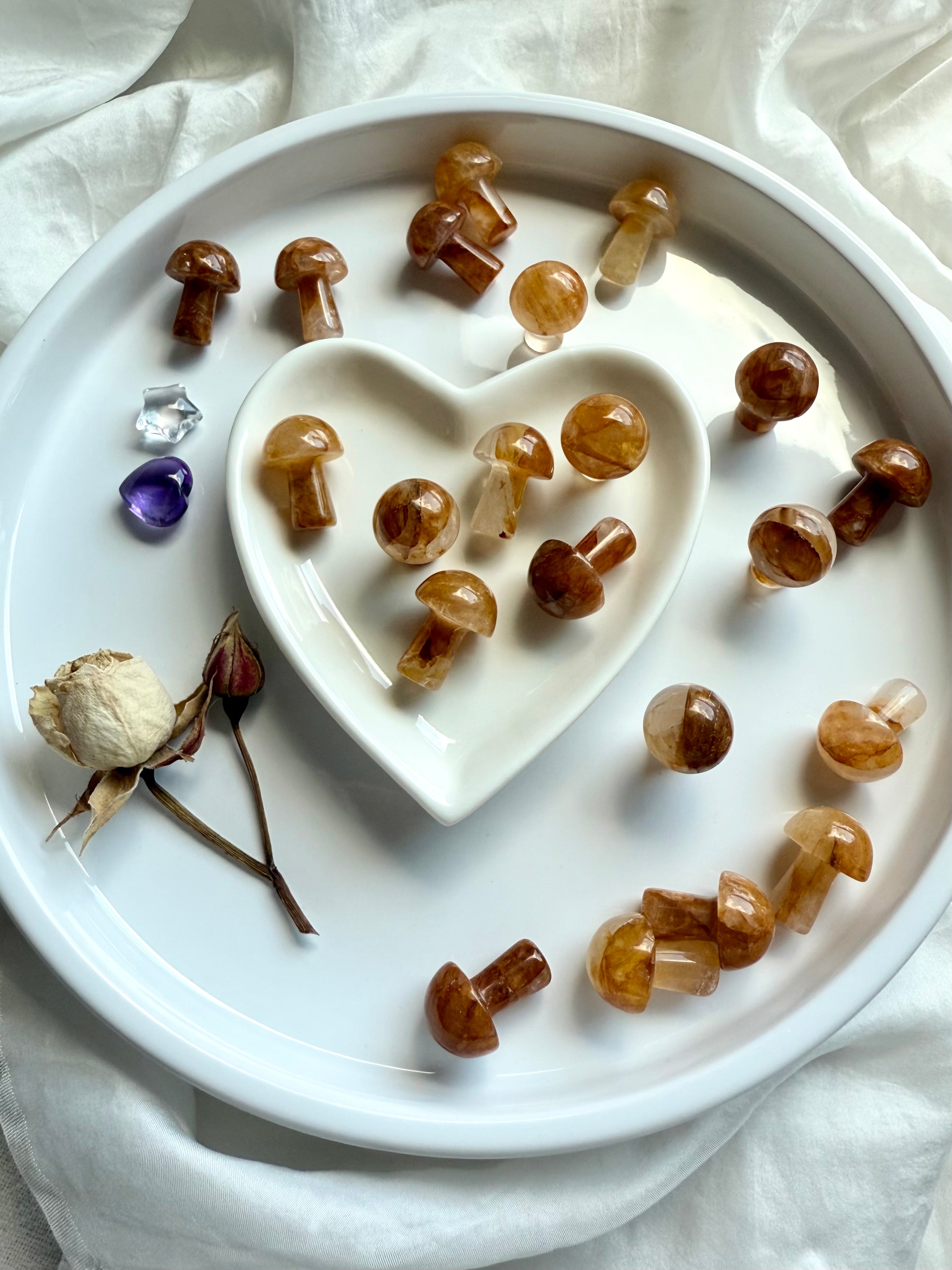 Golden Healer Quartz mushrooms arranged in white heart dish with dried rose and amethyst heart — whimsical crystal carvings for altar décor and gifting.