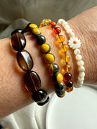 Stack of gemstone bracelets on wrist including Mother of Pearl daisy bracelet, bumblebee jasper, smoky quartz, and amber – crystal jewelry stack for lightness and vitality