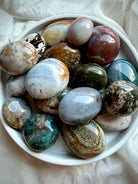Assorted Sea Jasper palm stones from Madagascar with green, cream, rust, and gray patterns displayed in a white ceramic bowl