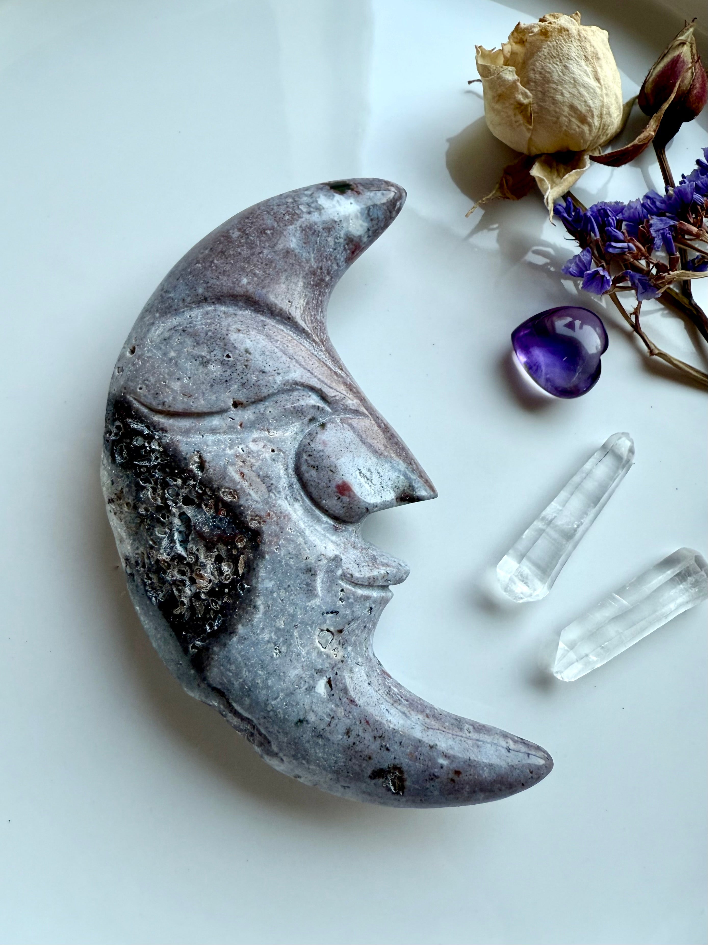 Purple Ocean Jasper moon face carving displayed with clear quartz points, amethyst heart, and flowers.