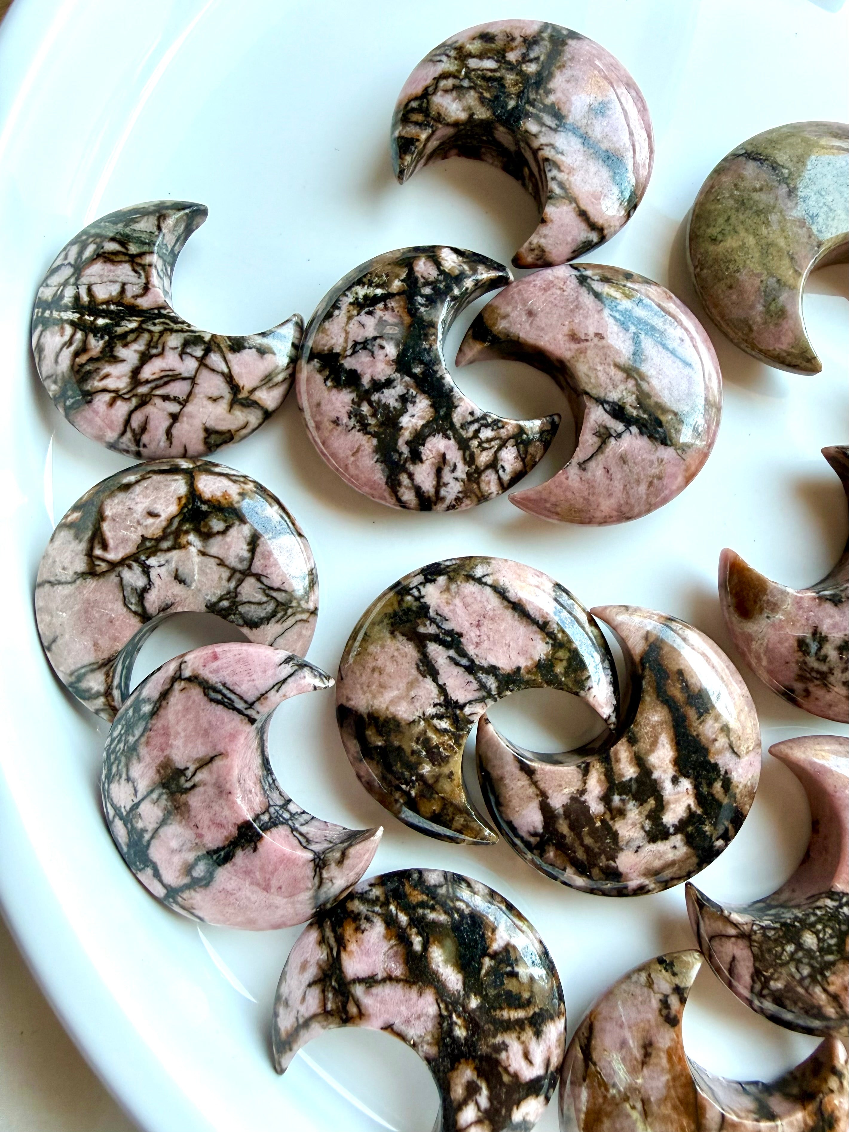Rhodonite moon carvings arranged on a white tray highlighting one-of-a-kind pink and black mineral patterns
