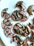 Rhodonite moon carvings arranged on a white tray highlighting one-of-a-kind pink and black mineral patterns