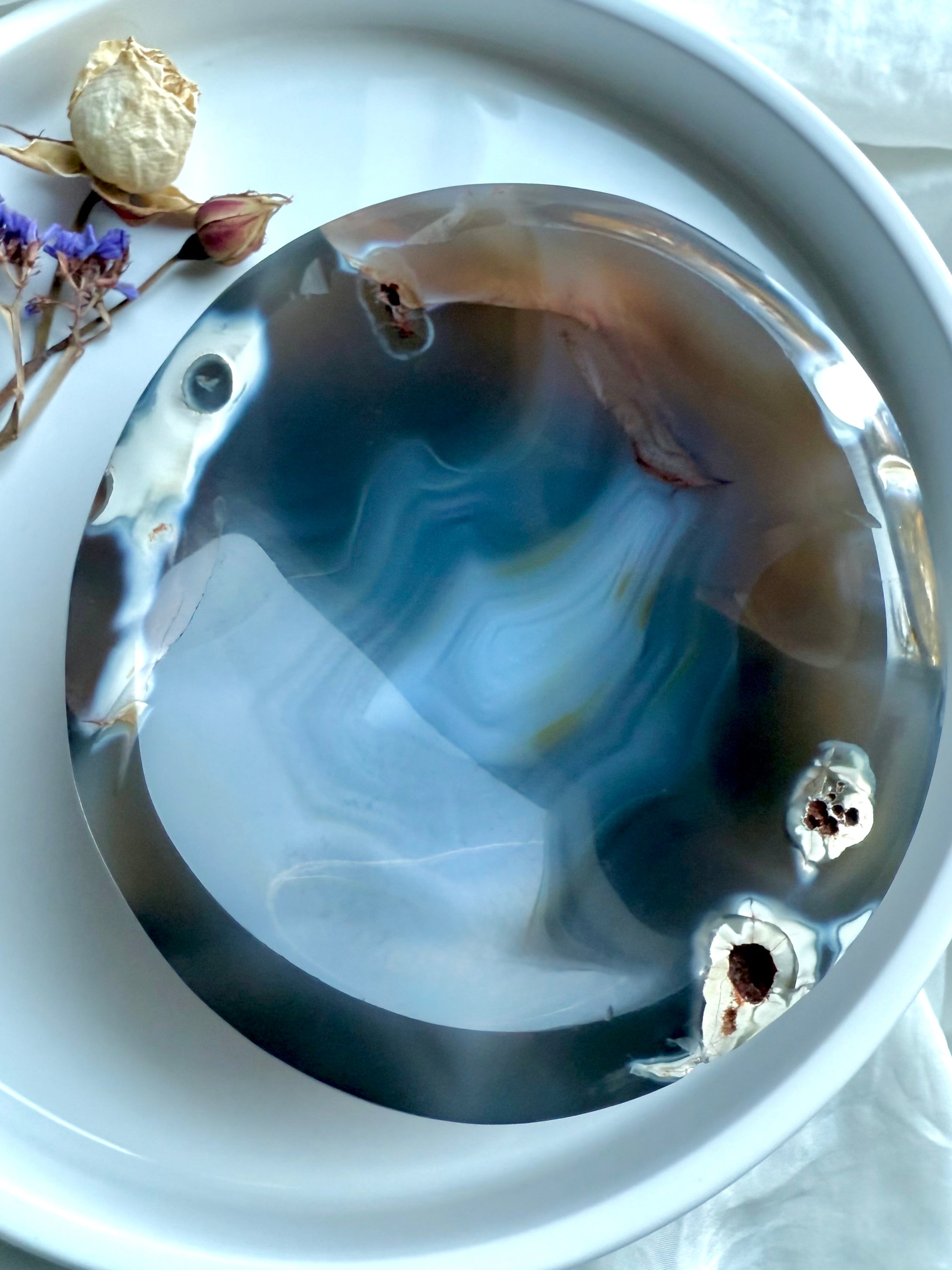 Overhead photo of polished Orca Agate crystal bowl on a white tray with dried flowers, selenite, and palo santo for cleansing rituals