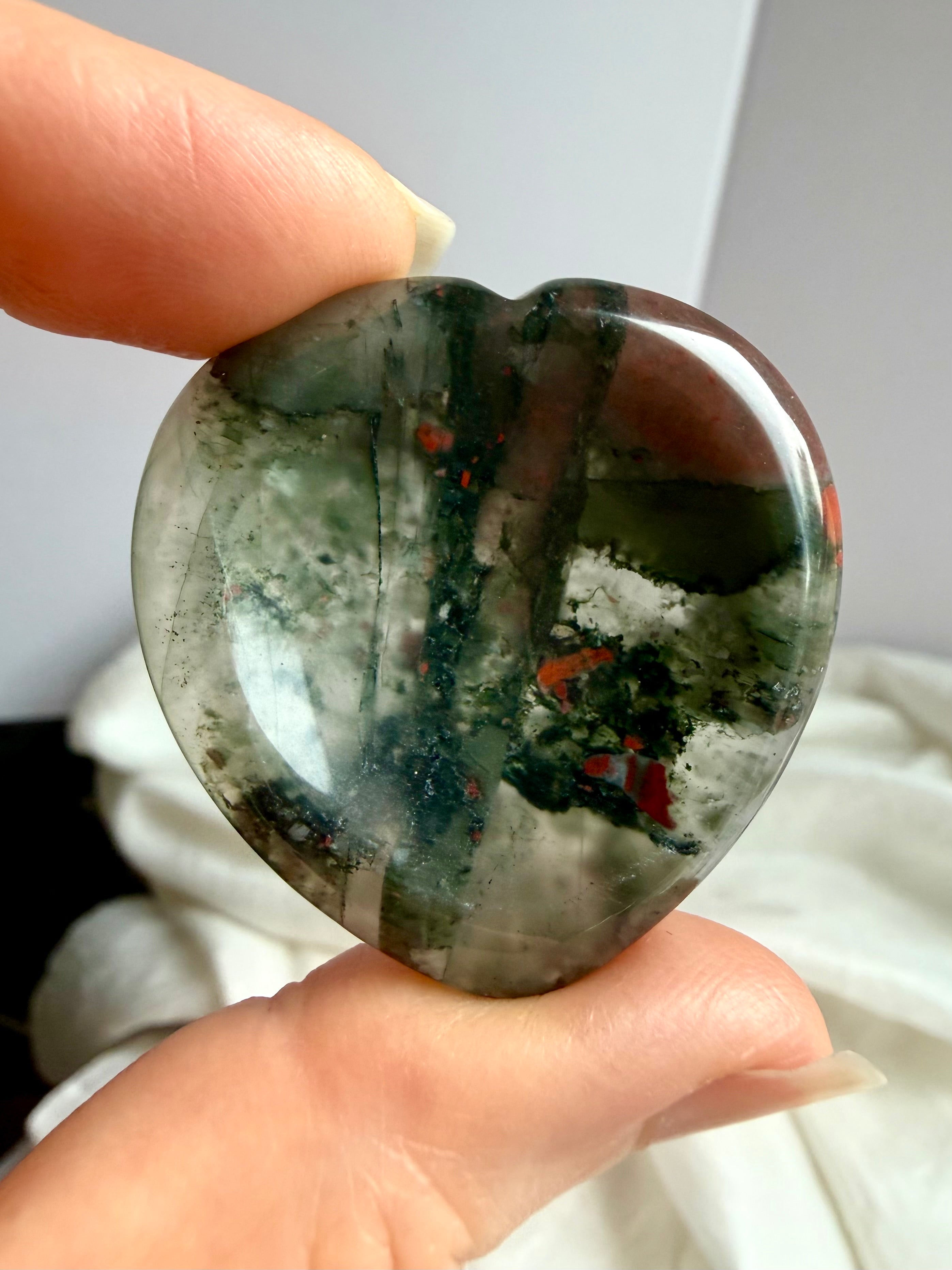 closeup hand holding bloodstone heart worry stone showing natural red flecks and polished finish, crystal for courage resilience and emotional balance