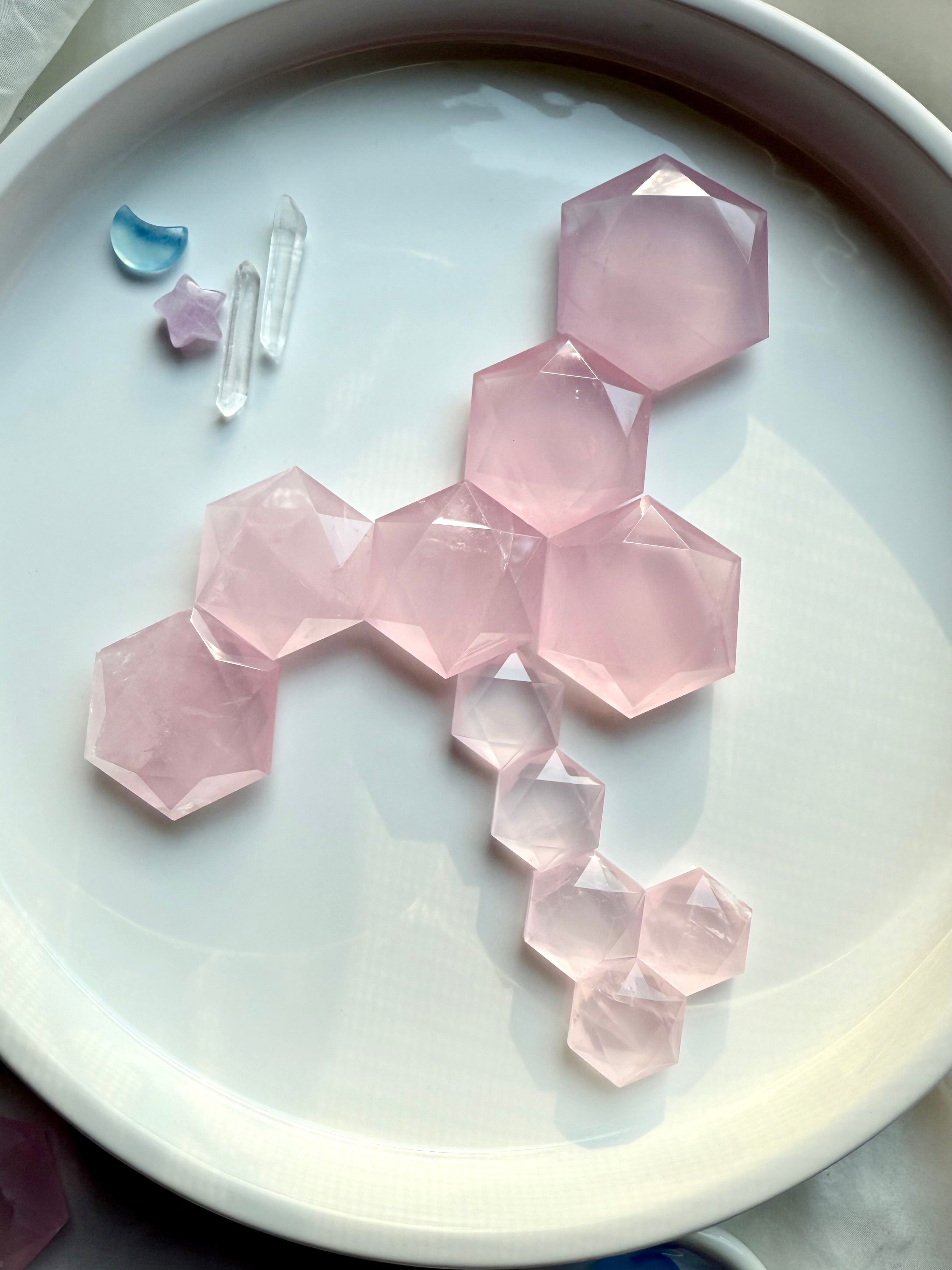 Faceted rose quartz hexagon crystals arranged by size on a white tray, with mini moon, star, and clear quartz accents for heart chakra healing and crystal grid work.