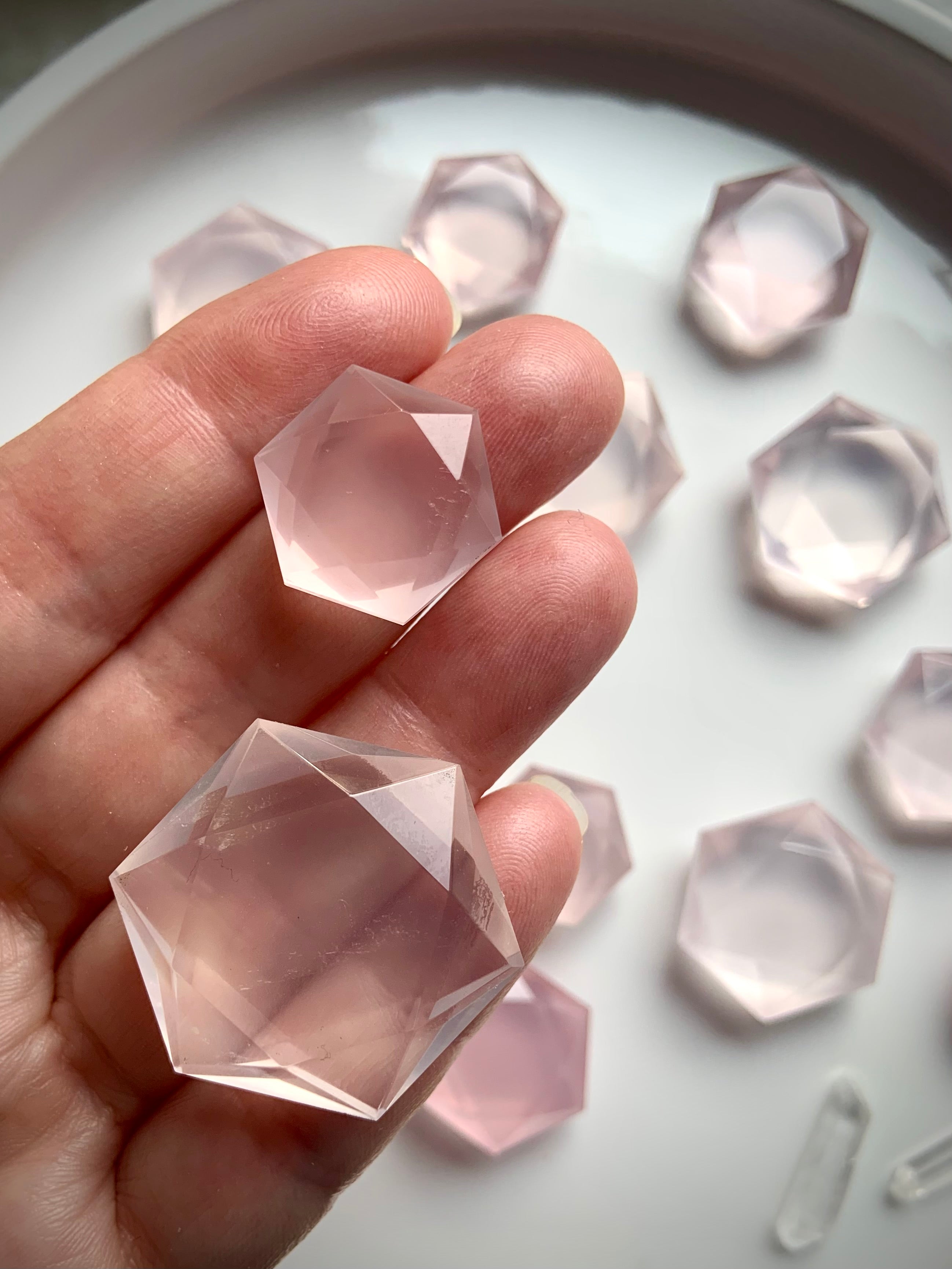 Hand holding smallest and largest faceted Rose Quartz hexagon crystals, translucent pink sacred geometry gems with plate of crystals in background