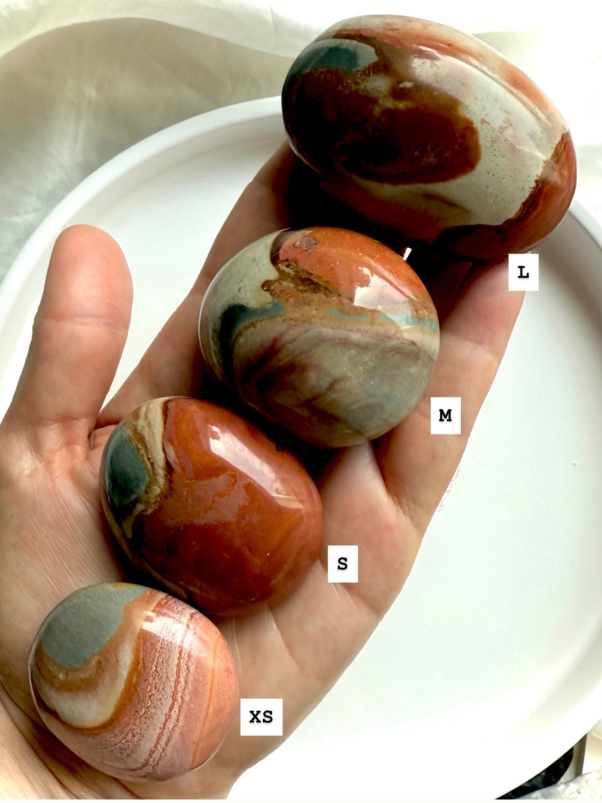Three sizes of Polychrome Jasper palm stones shown in hand, labeled XS, S, M, and L