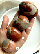 Three sizes of Polychrome Jasper palm stones shown in hand, labeled XS, S, M, and L