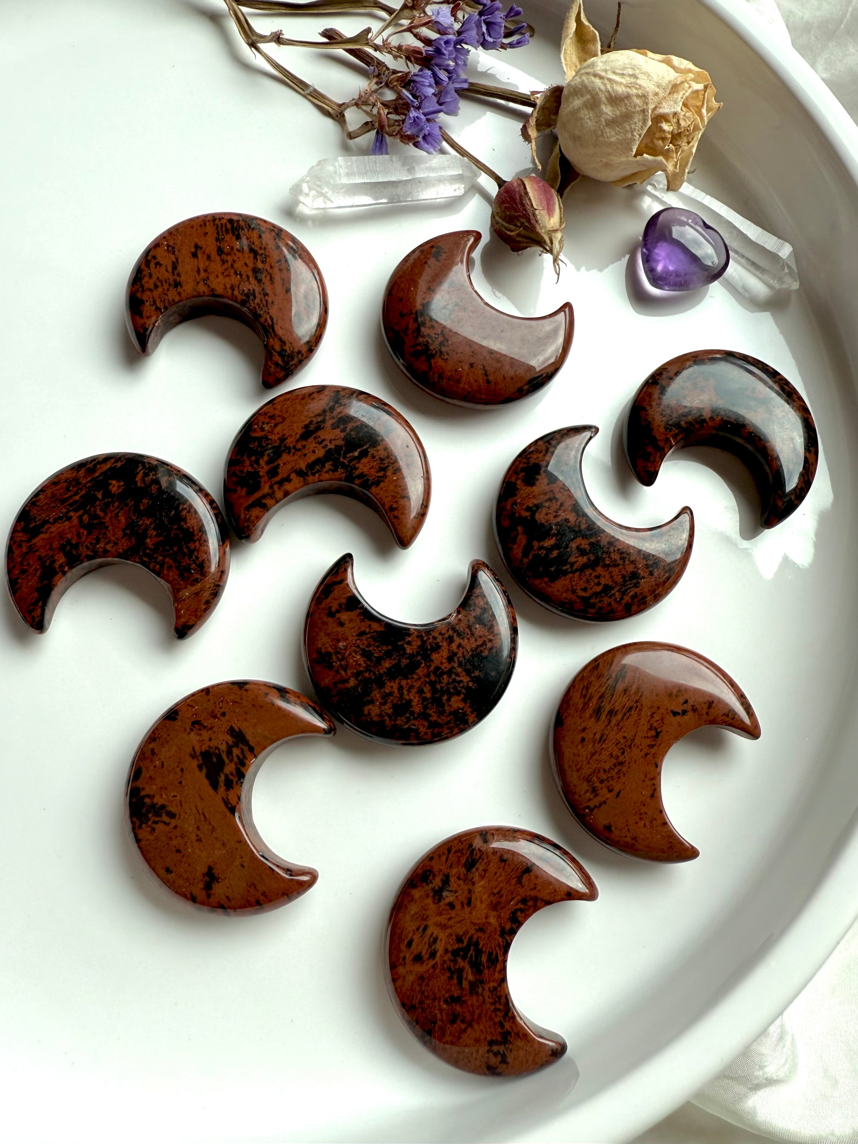 Mahogany Obsidian crescent moons on white plate with flowers & crystals. Grounding Root Chakra carvings for energetic protection.
