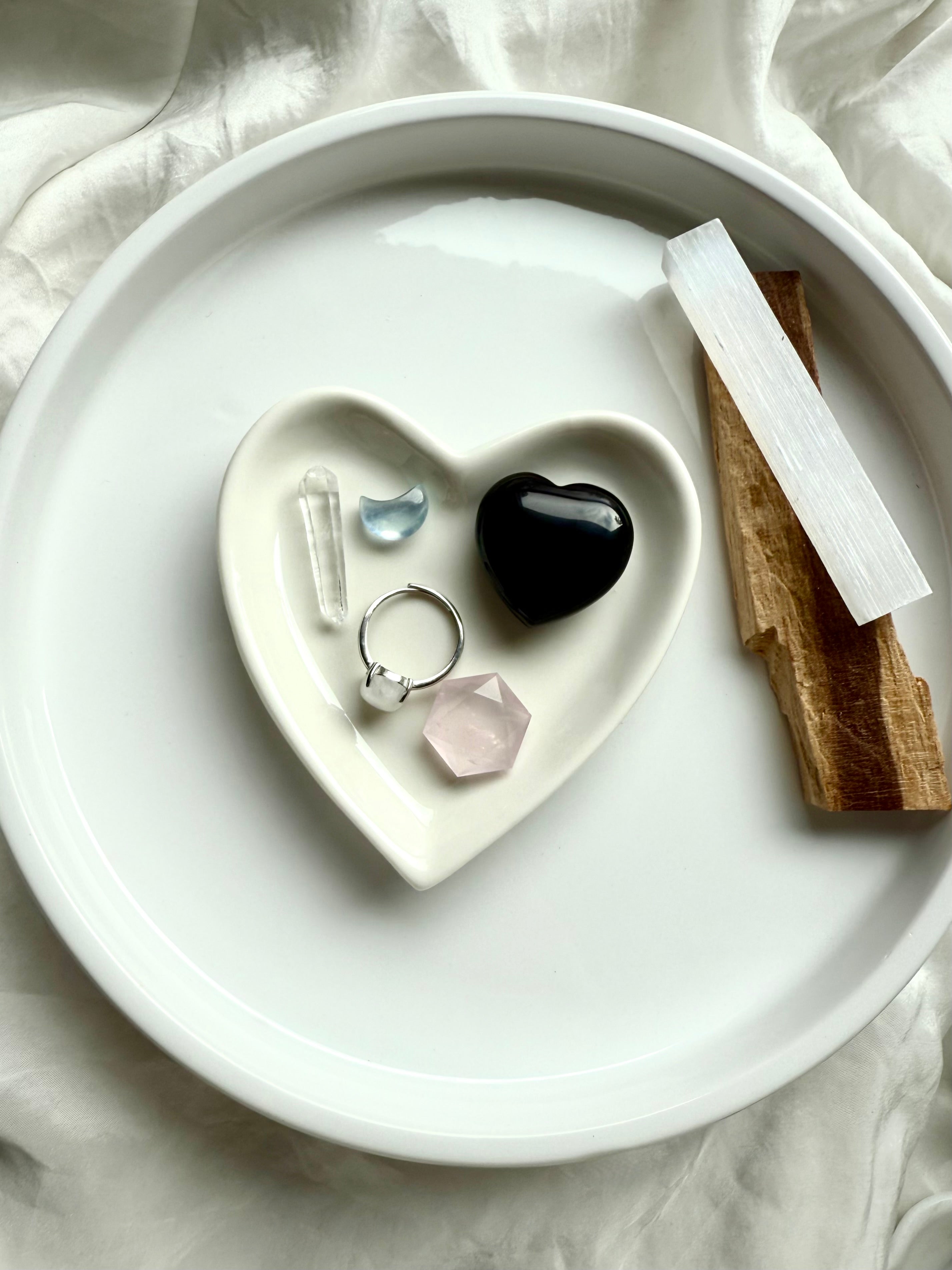 Black Obsidian heart in a white ceramic dish with other small crystals, jewelry, and palo santo.