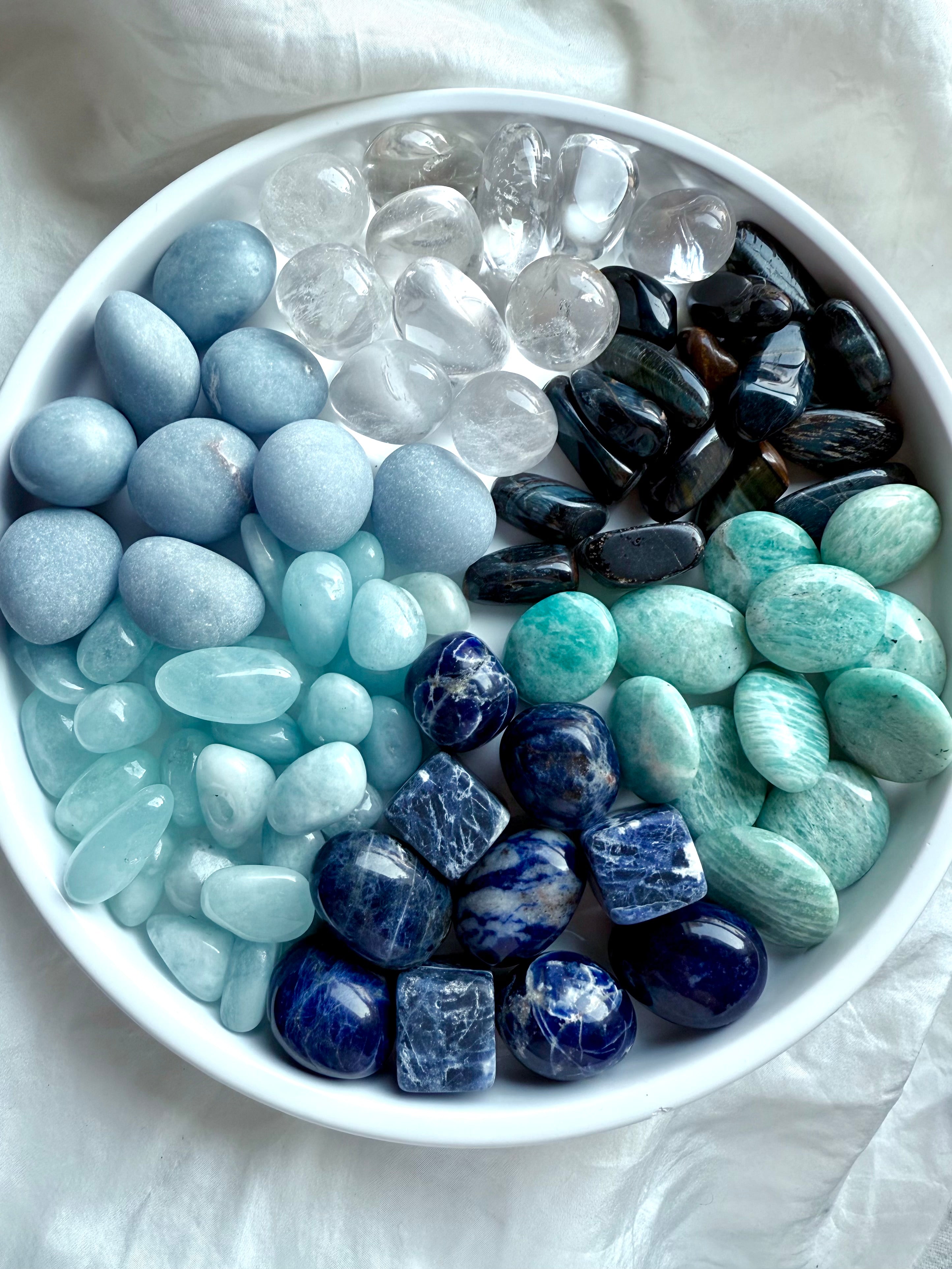 Bowl of throat chakra crystals — Angelite, Sodalite, Amazonite, Aquamarine, Blue Tiger’s Eye, and Clear Quartz healing stones