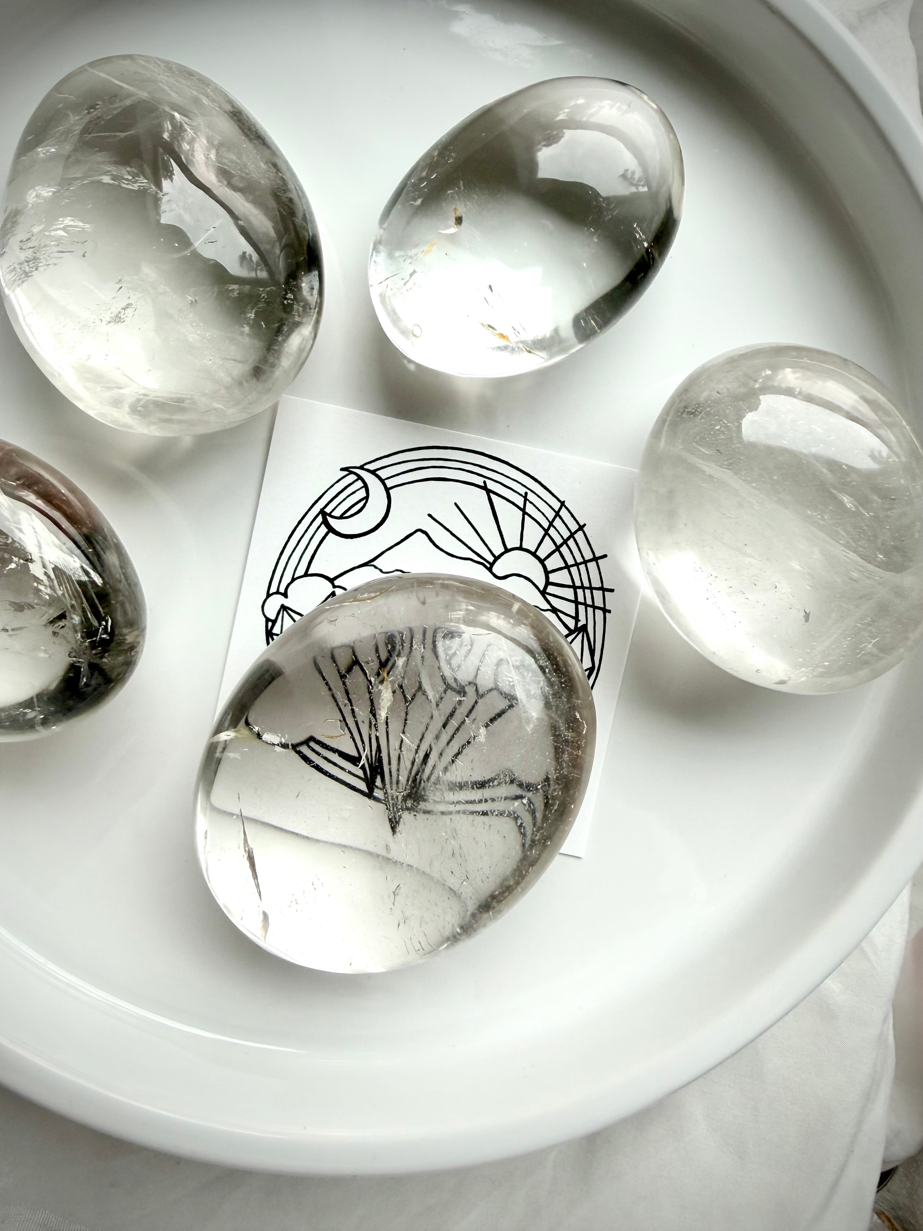 clear quartz palm stones on white tray with brand card beneath stone showing high clarity and natural inclusions