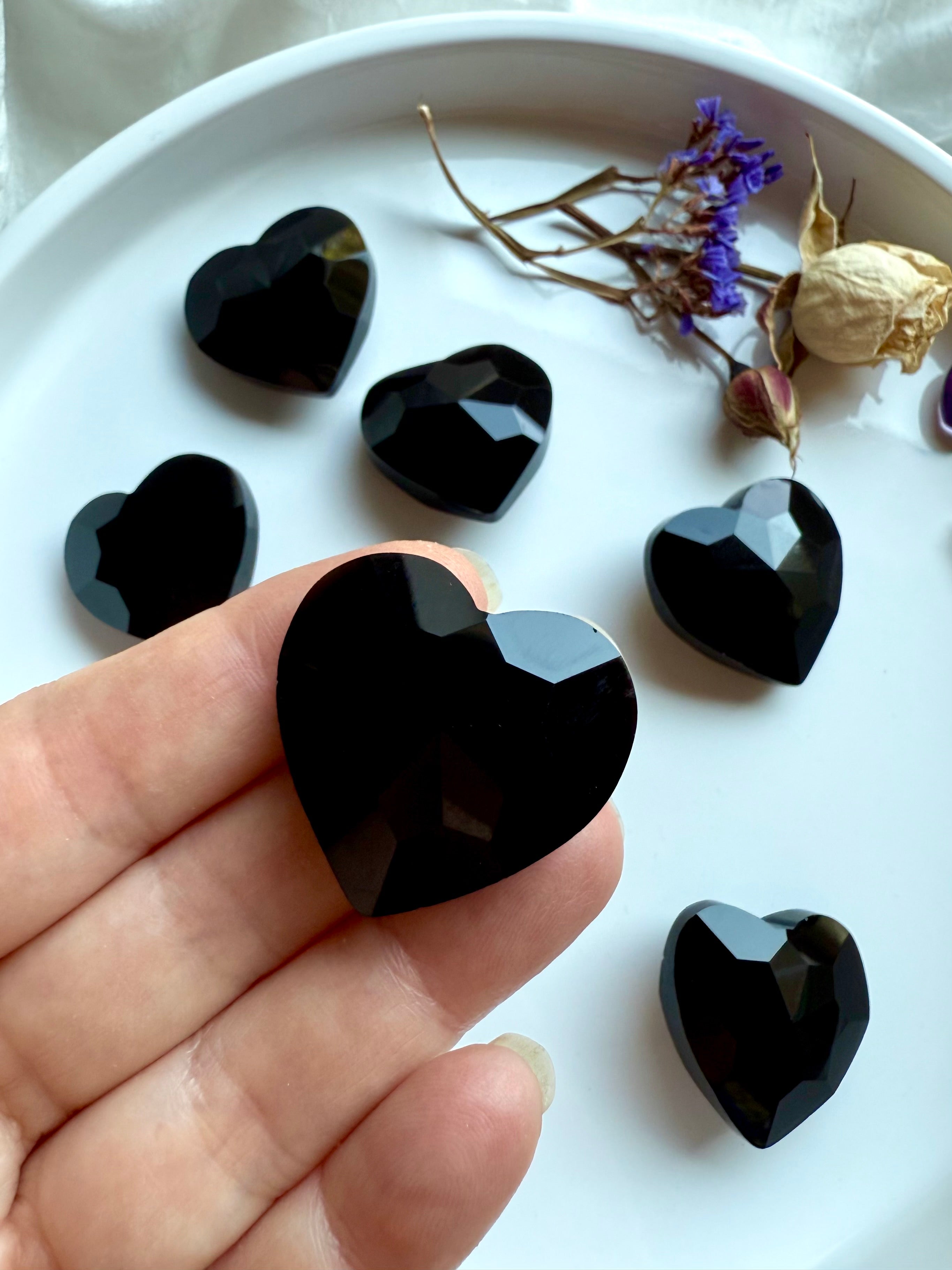 Closeup of black obsidian heart-shaped gems on fingertips for scale with more in the background on a white plate with dried flowers