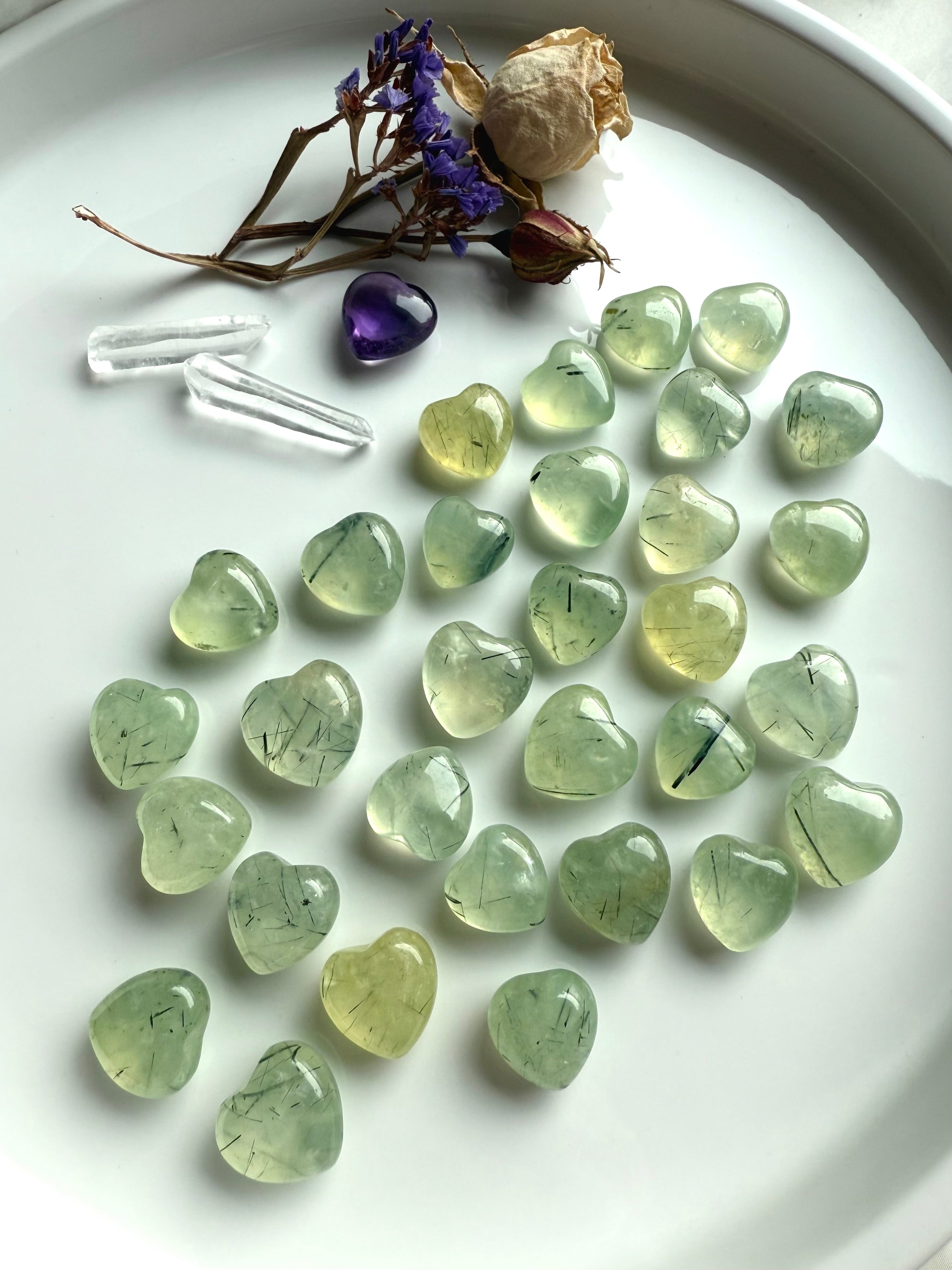 Mini Prehnite crystal hearts with black tourmaline inclusions, displayed on a white tray with dried flowers and quartz points.