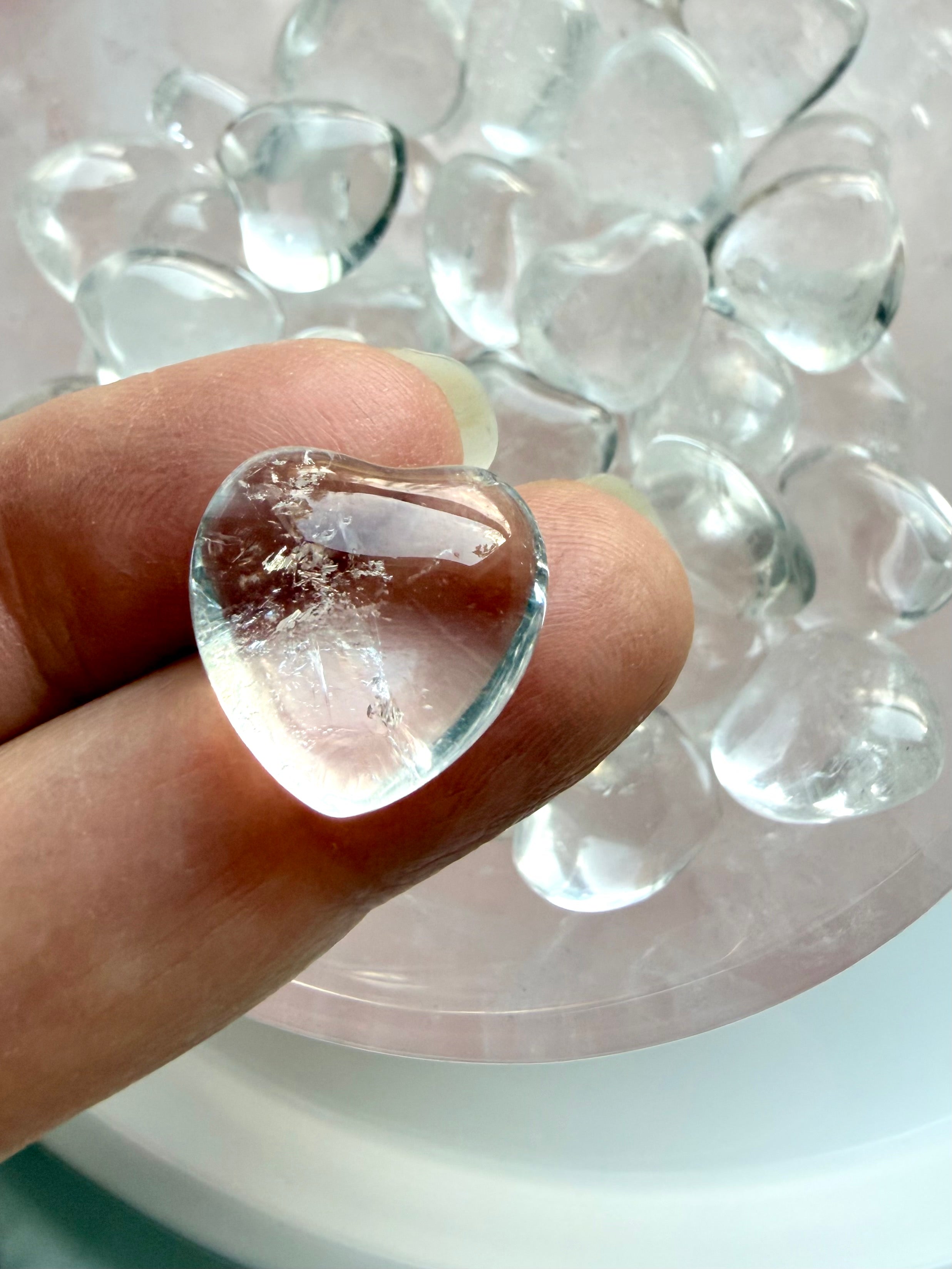 Mini Clear Quartz heart crystal on fingertip showing high clarity quartz with bowl of hearts in background