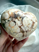 Hand holding a Flower Agate heart at an angle, capturing its blooming patterns and soft pastel hues — gentle crystal for emotional balance, resilience, and renewal.